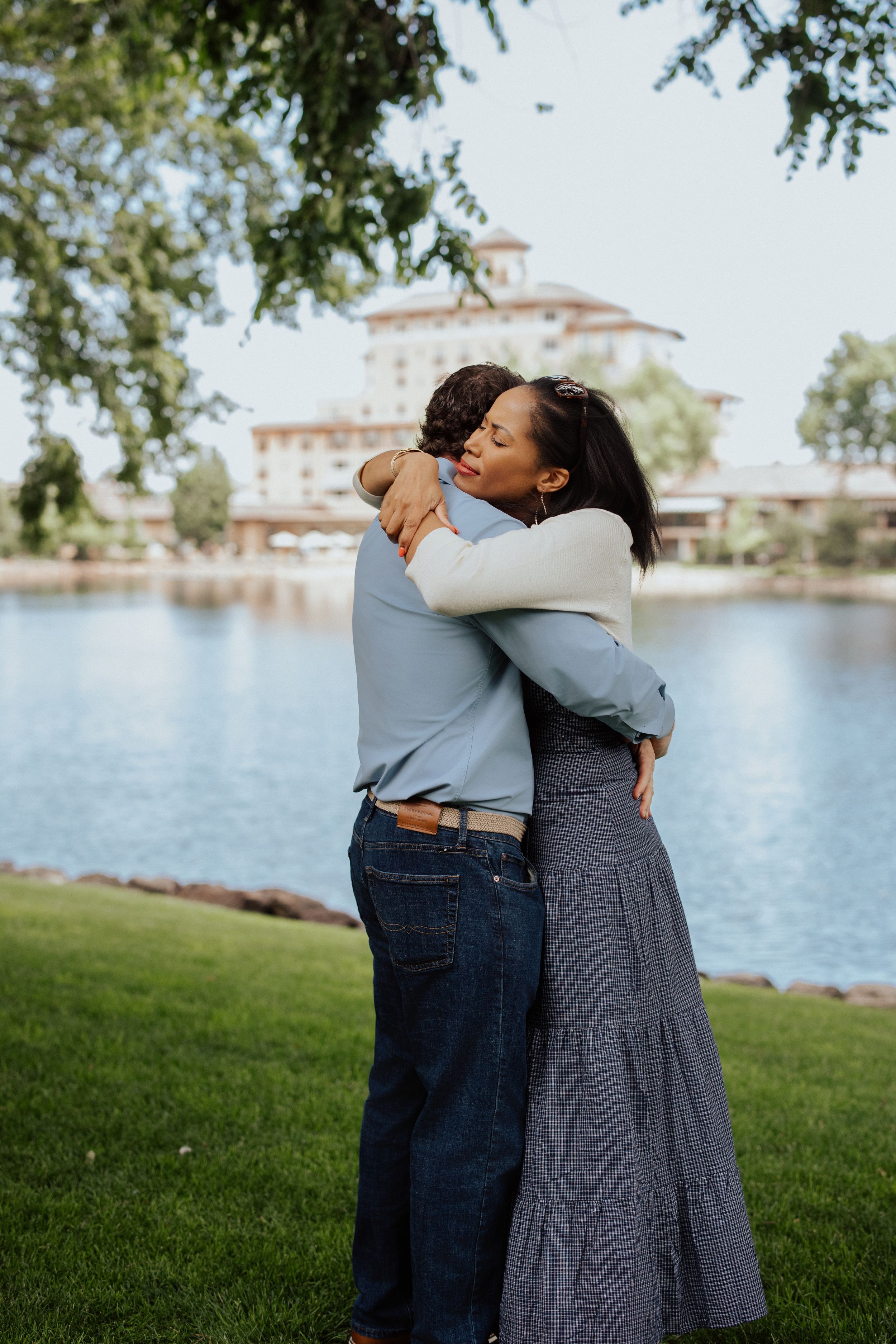 A couple shares a tender embrace on a grassy lawn beside a lake with a building in the background.