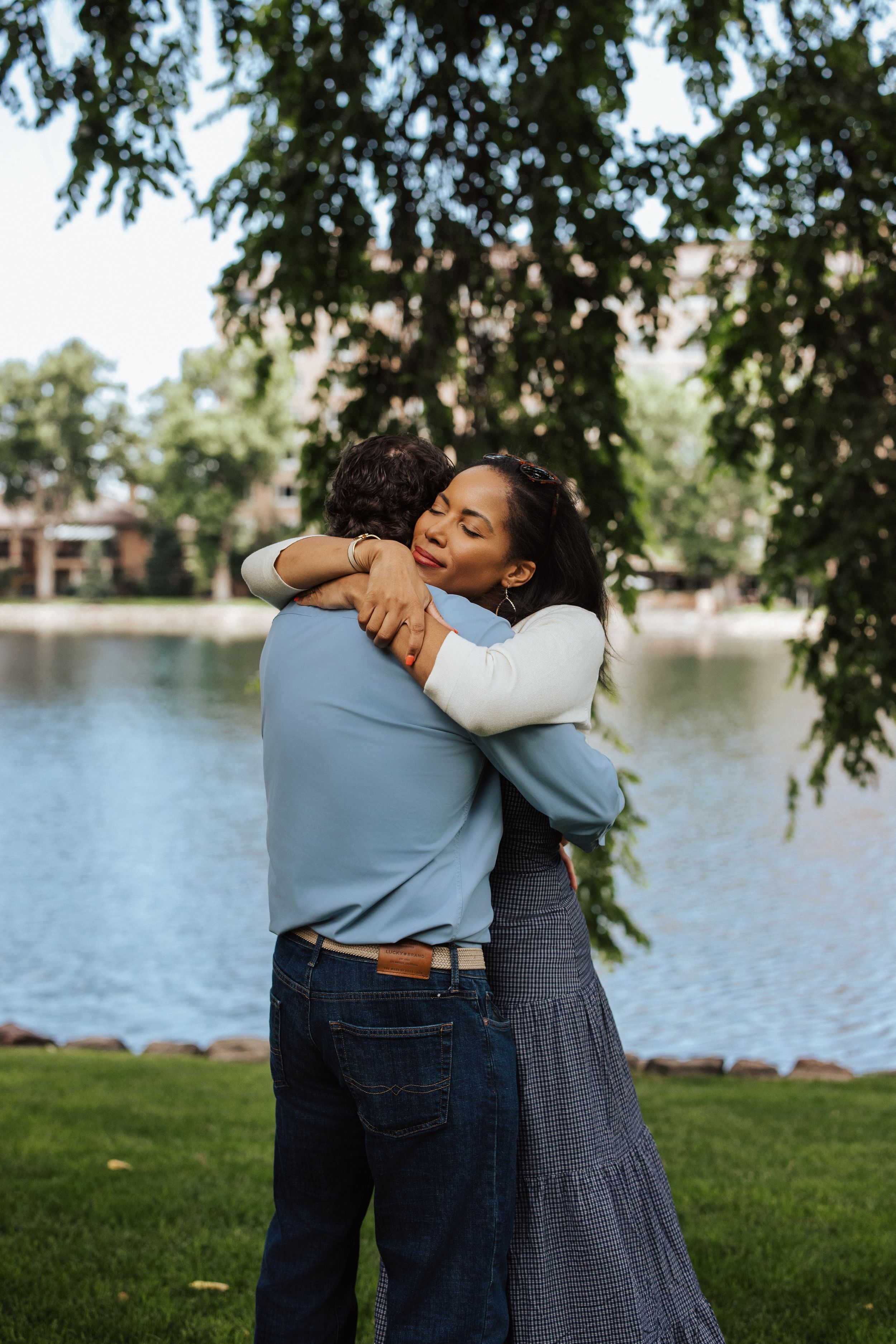 A couple shares a tender embrace by a serene lake on a sunny day, with tree branches framing the scene.