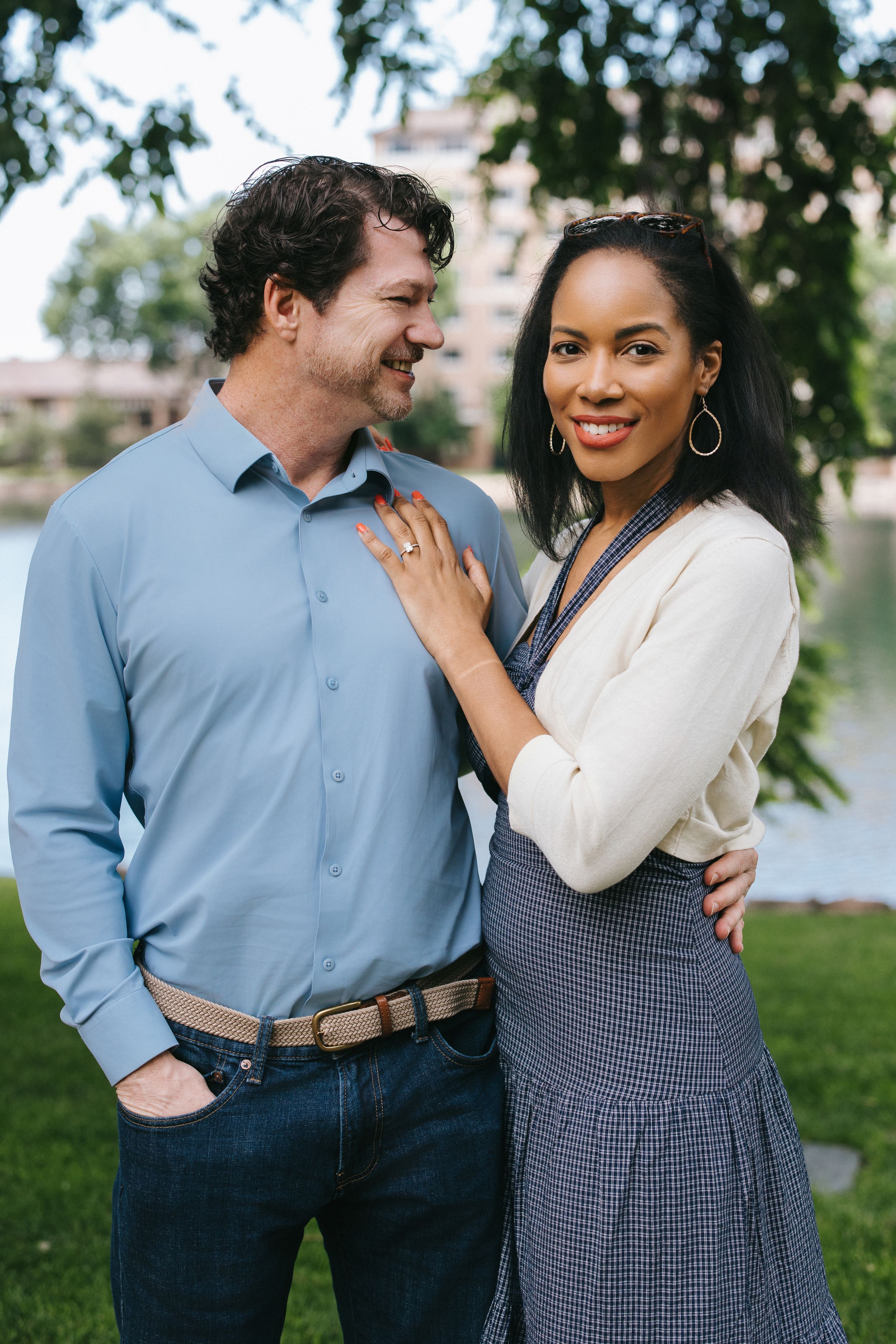 A couple smiles at each other outdoors, wearing coordinated business casual attire against a natural background.