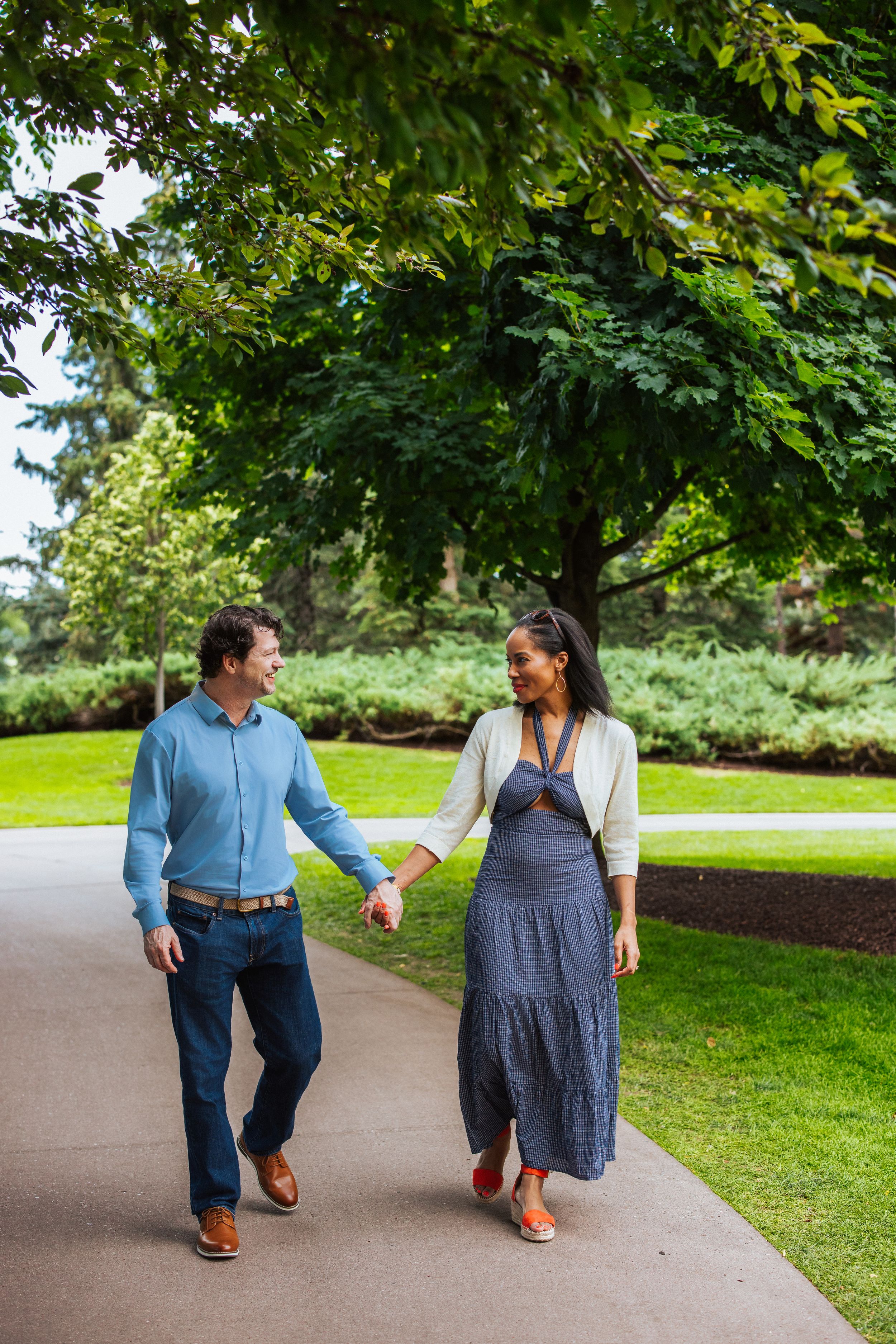 A couple walks together on a garden path surrounded by lush green trees and landscaping.