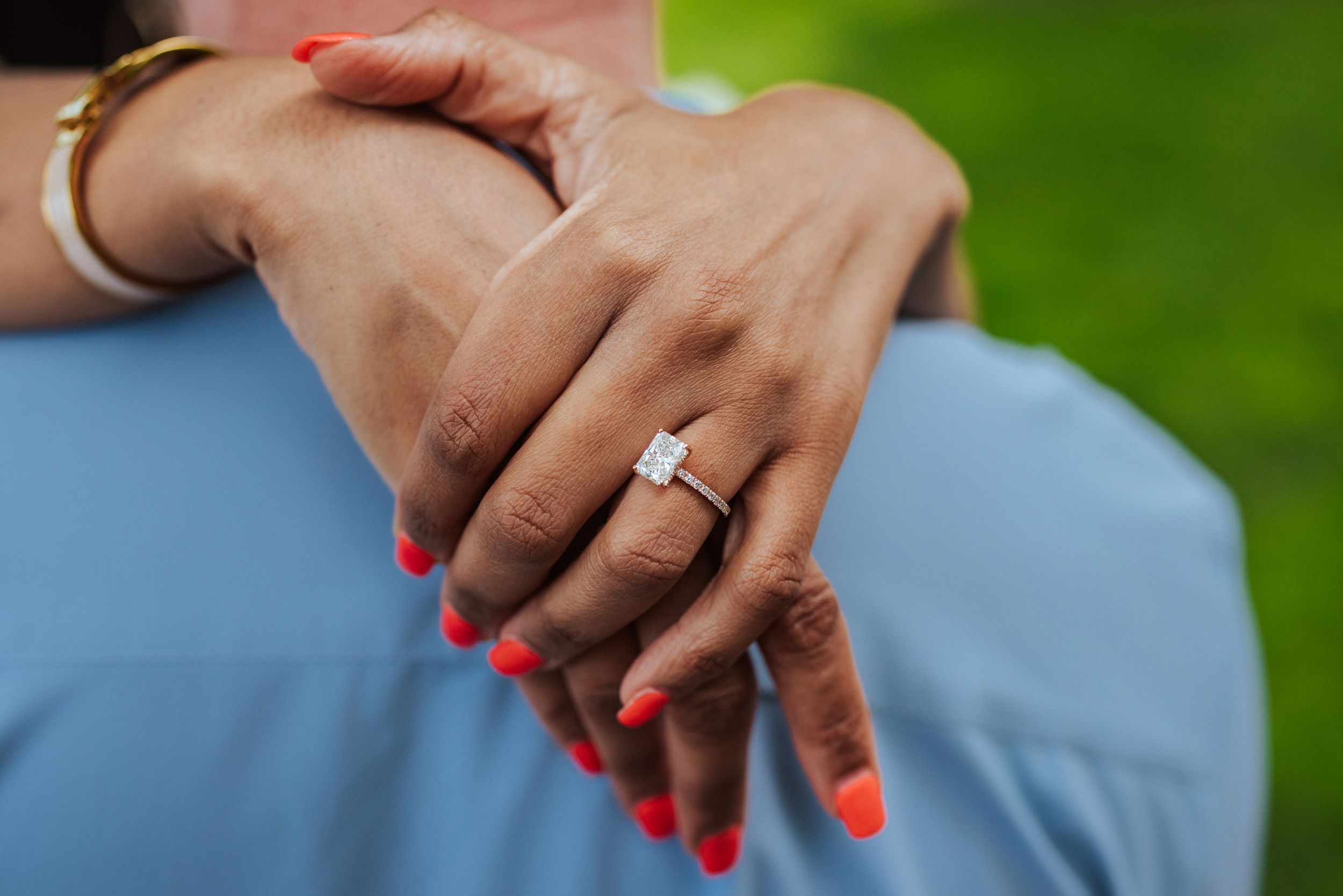 Hands with bright red nail polish hold a white engagement ring against a blurred green background.