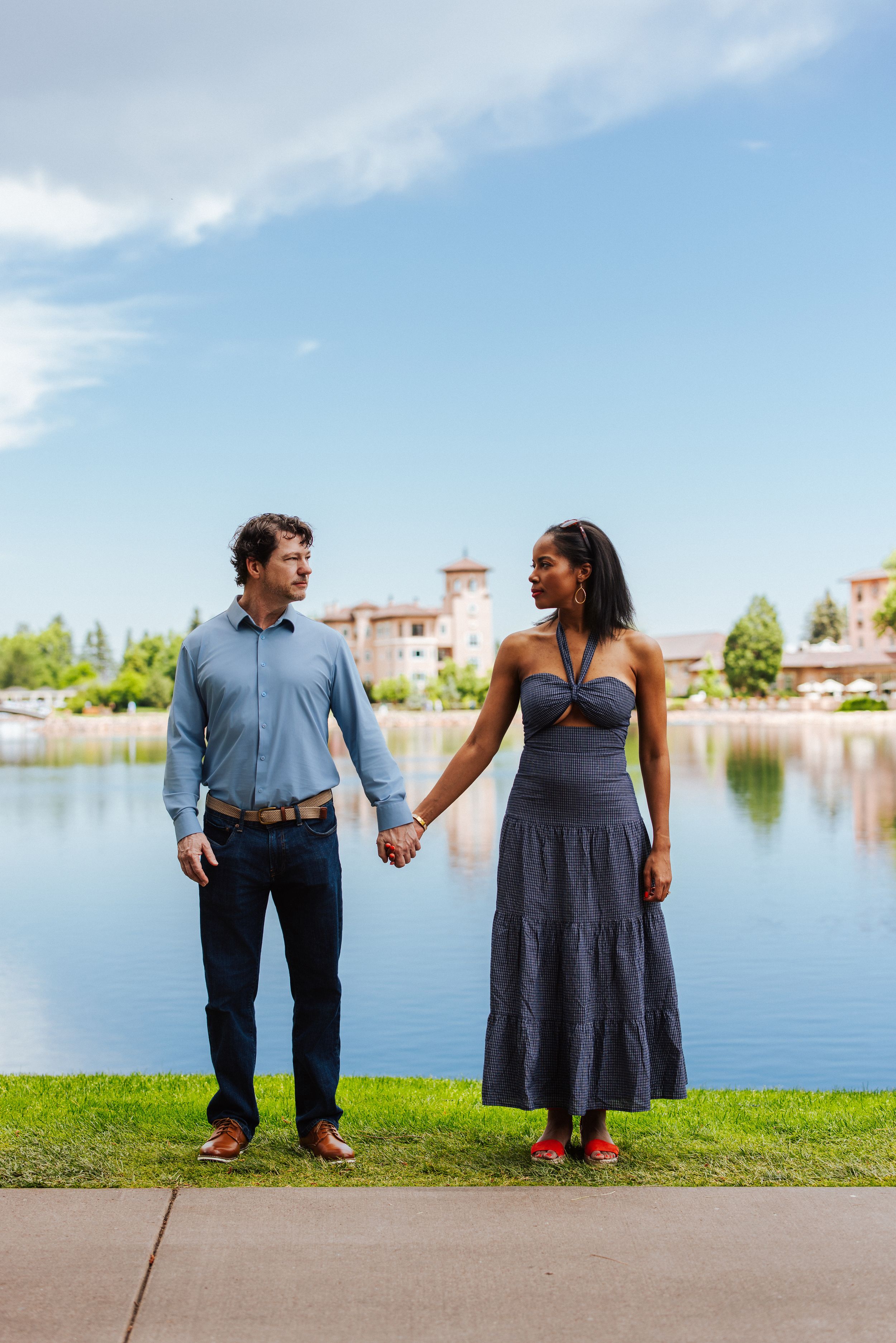 A couple holds hands while standing on a path beside a scenic lake with Mediterranean-style buildings in the background.