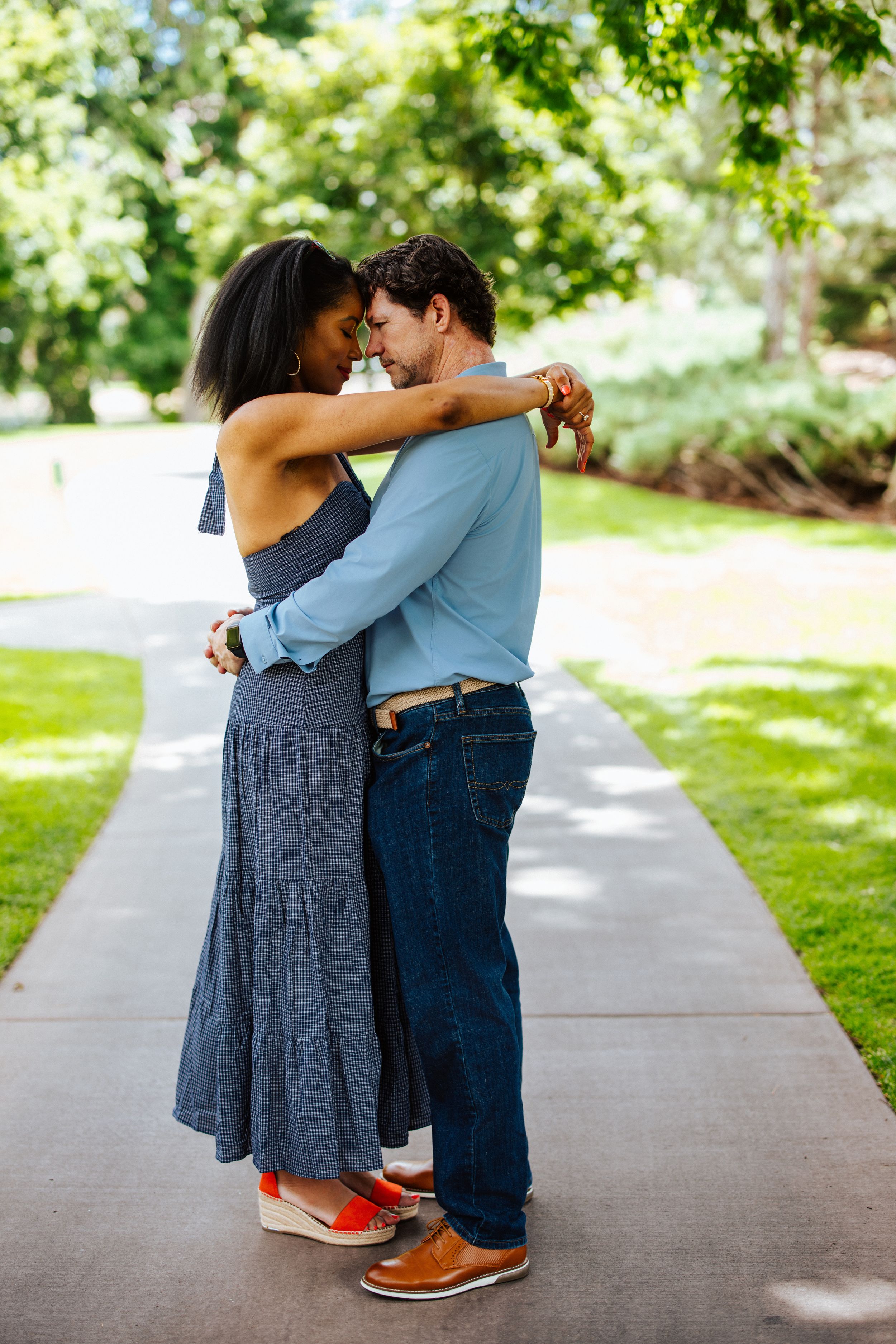 A couple embraces on a sunny pathway surrounded by green trees and lawn, standing close in casual summer outfits.