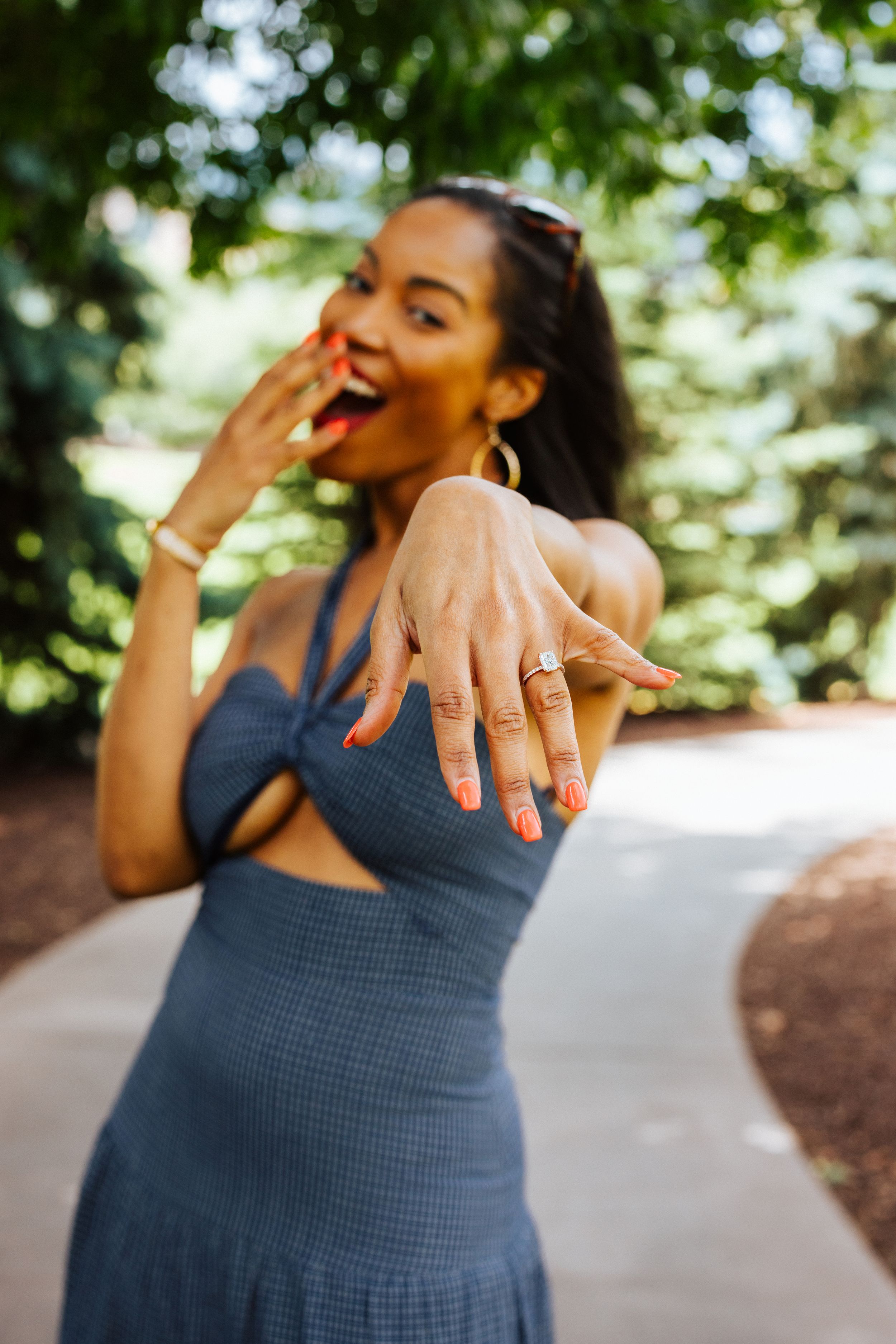 A joyful woman in a blue dress shows off her engagement ring while laughing in a garden setting.