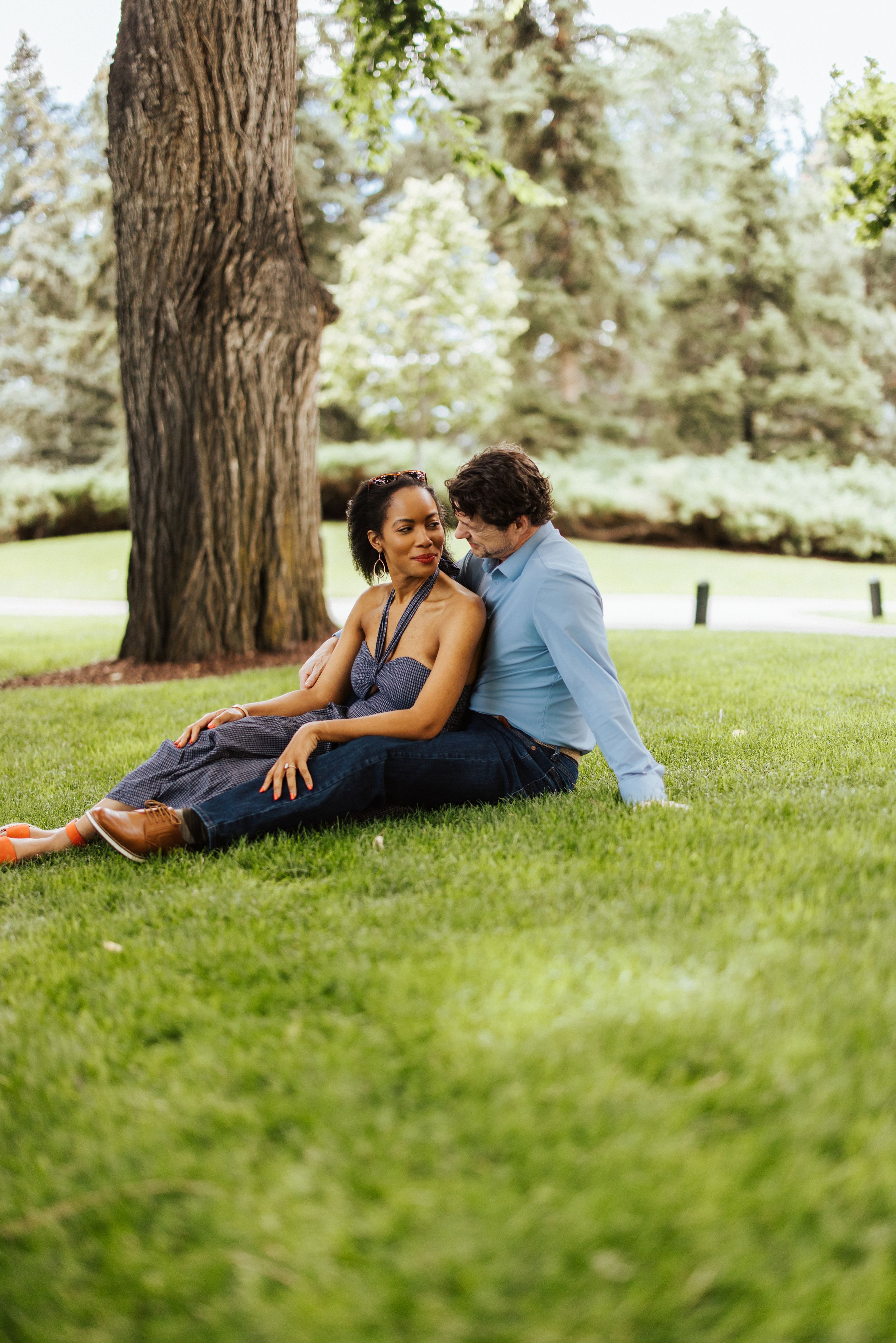 A couple relaxes together lying on green grass near a tree trunk in a sunny park setting.