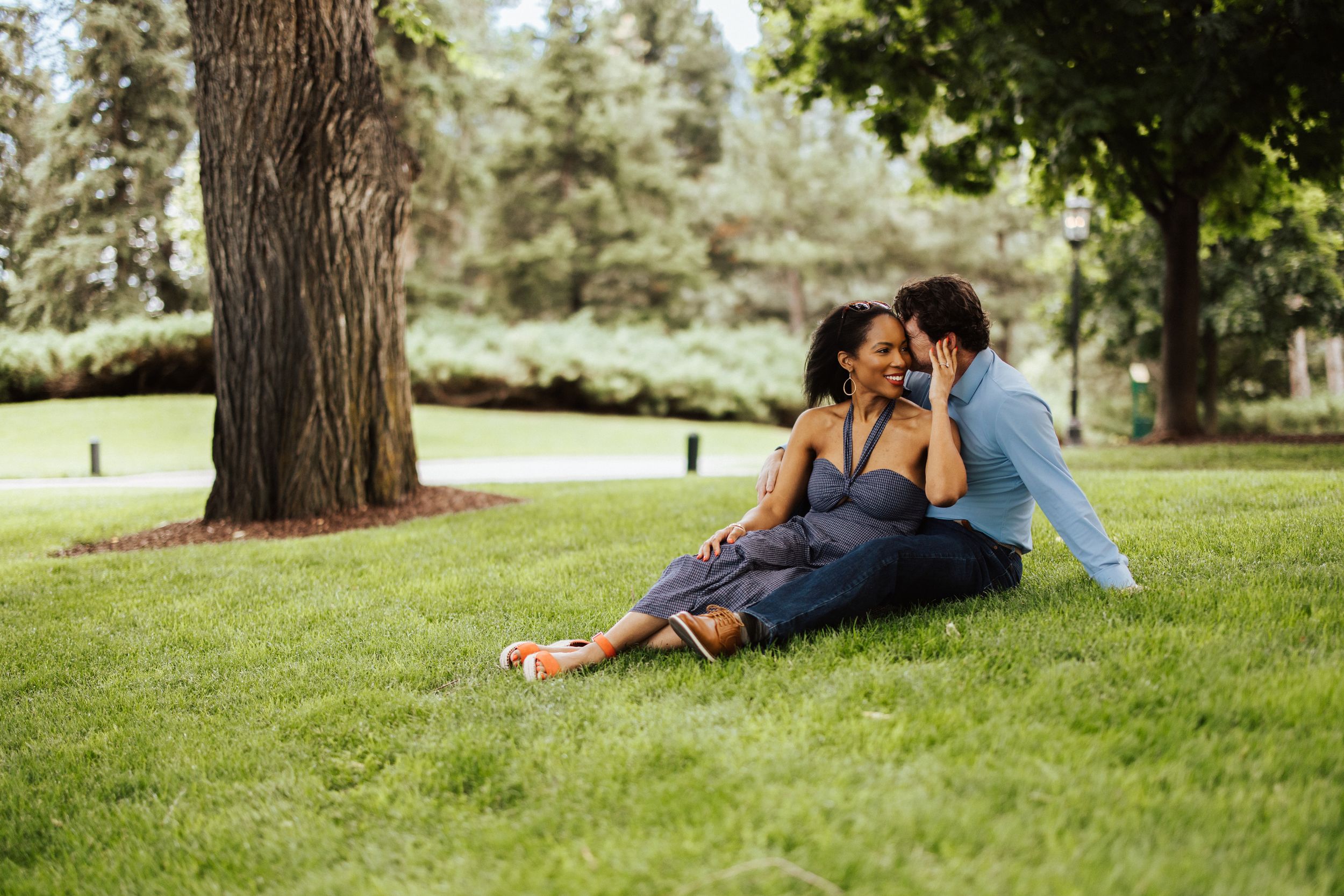 Two people relax under the shade of a tree on a grassy slope during a sunny afternoon.