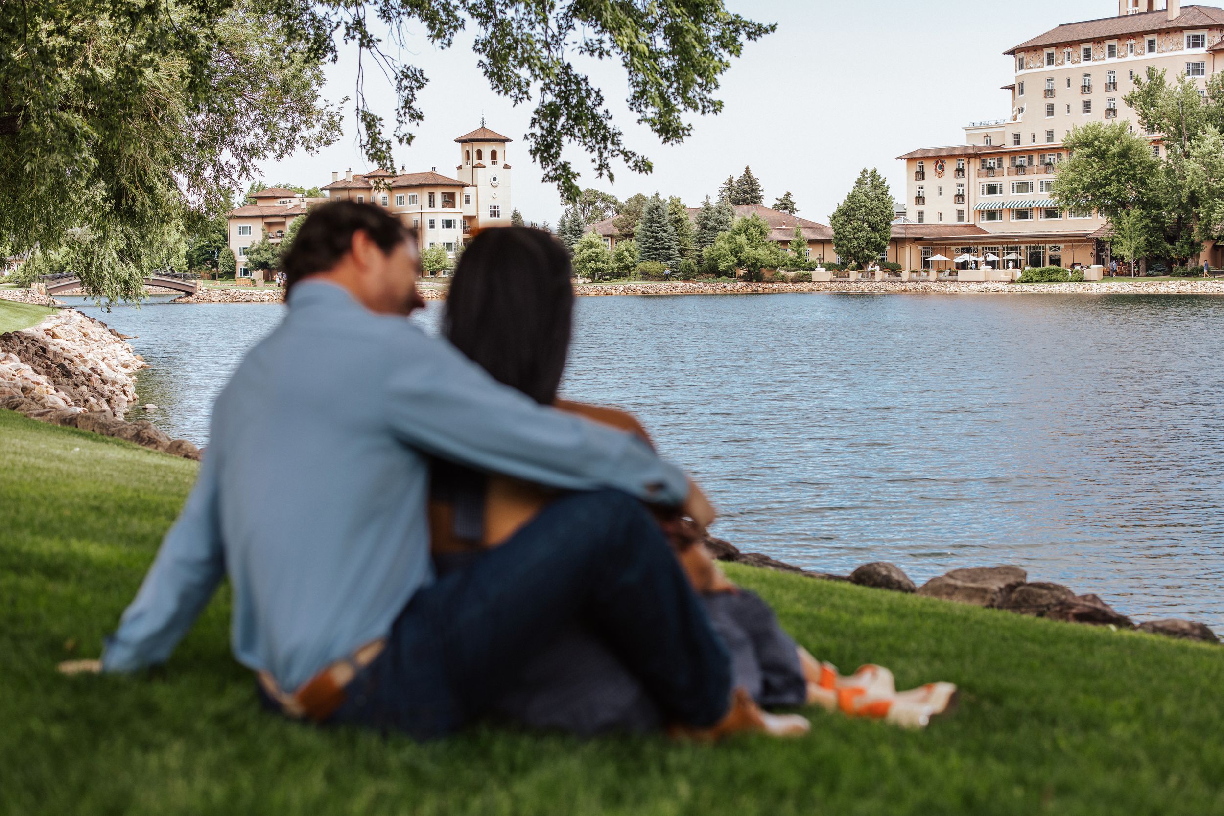 A couple sits together on a grassy lakeside overlooking a resort building on a sunny afternoon.