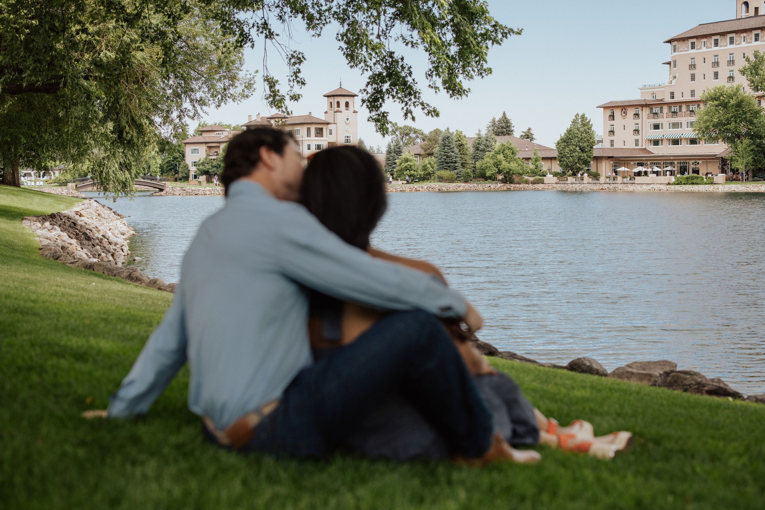 Couple enjoys romantic lakeside moment while sitting on grassy shore with historic buildings in background.