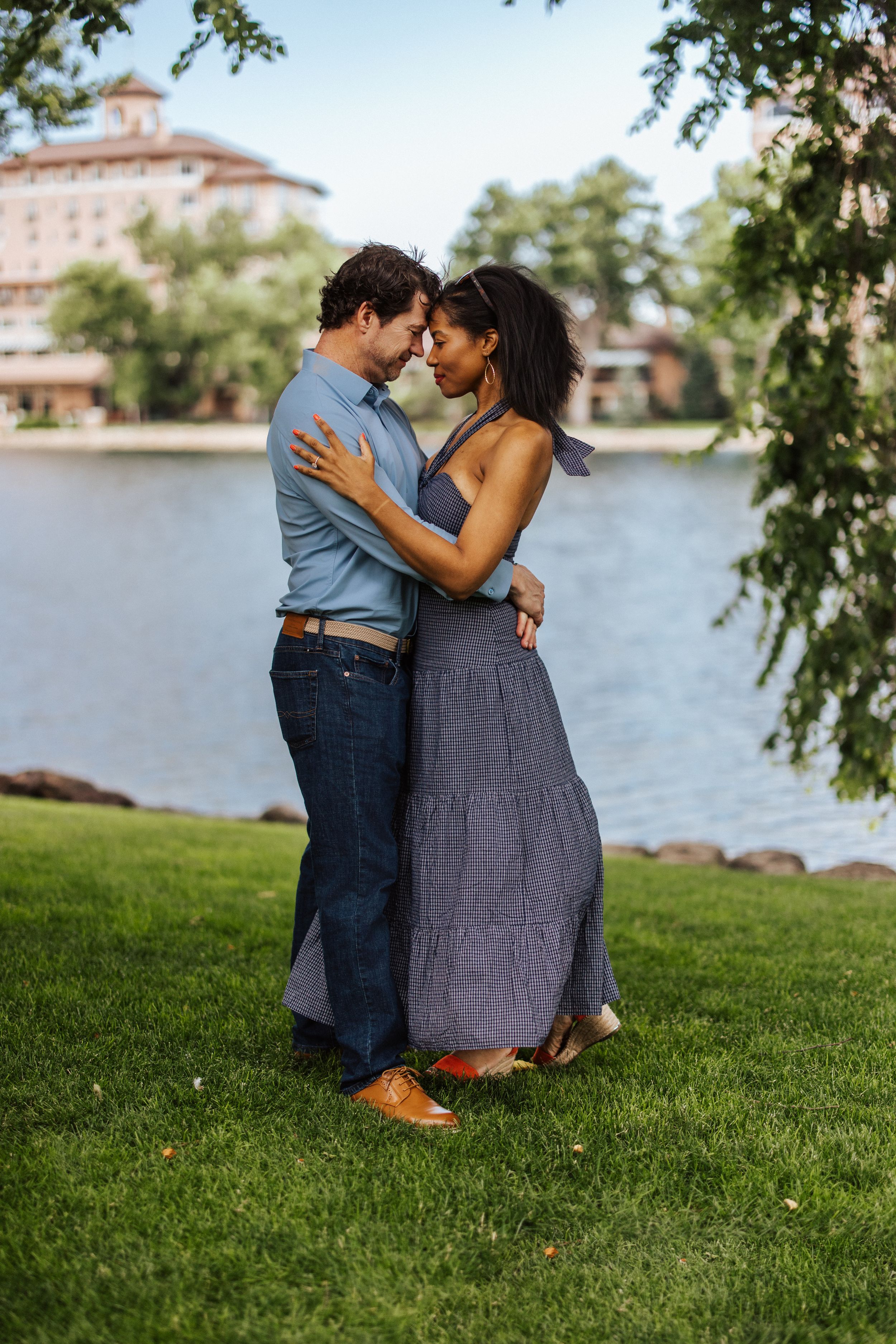A couple shares an intimate moment while embracing on a grassy lawn beside a lake with a grand building in the background.