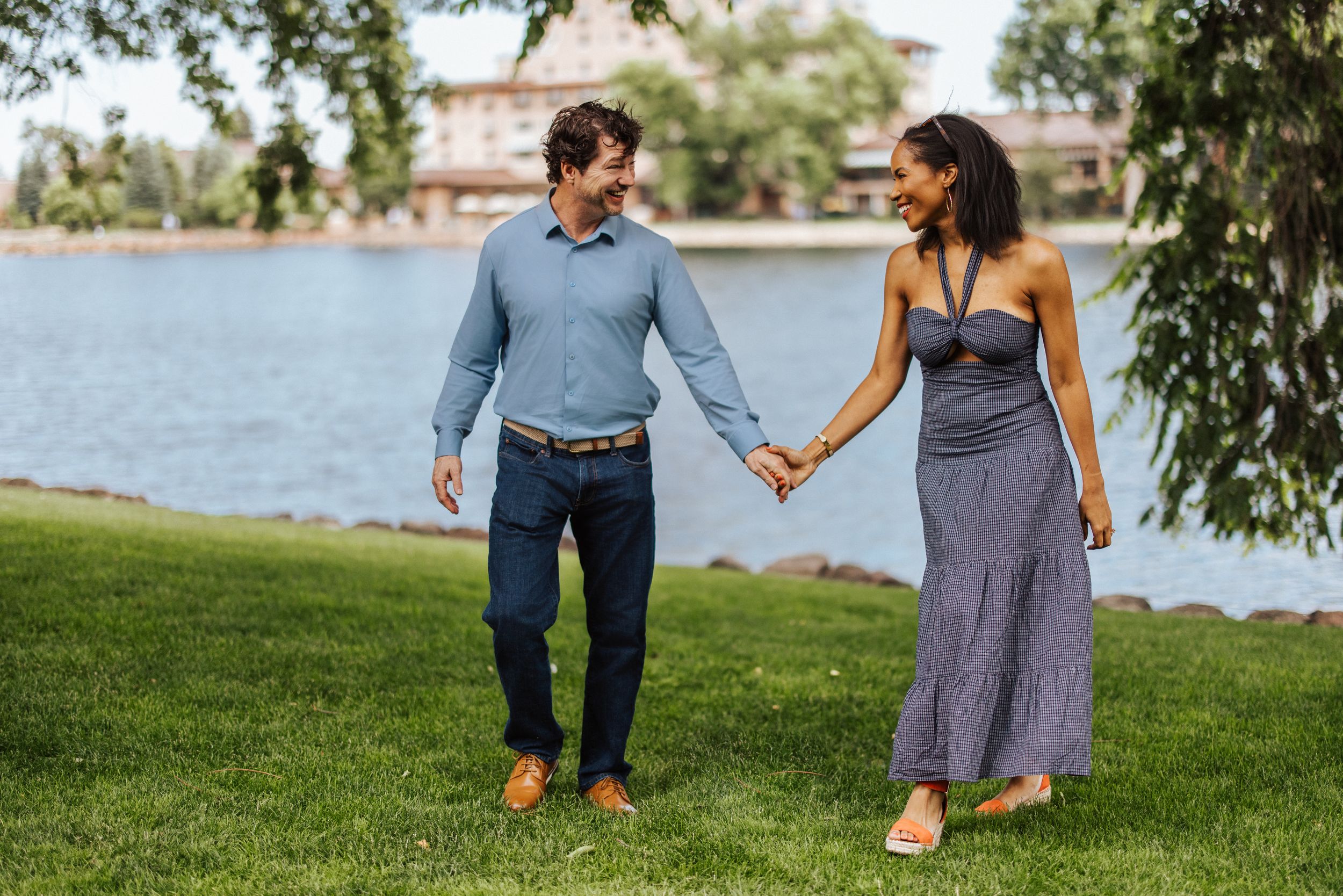 A couple walks hand in hand along a grassy lake shore on a sunny summer day.