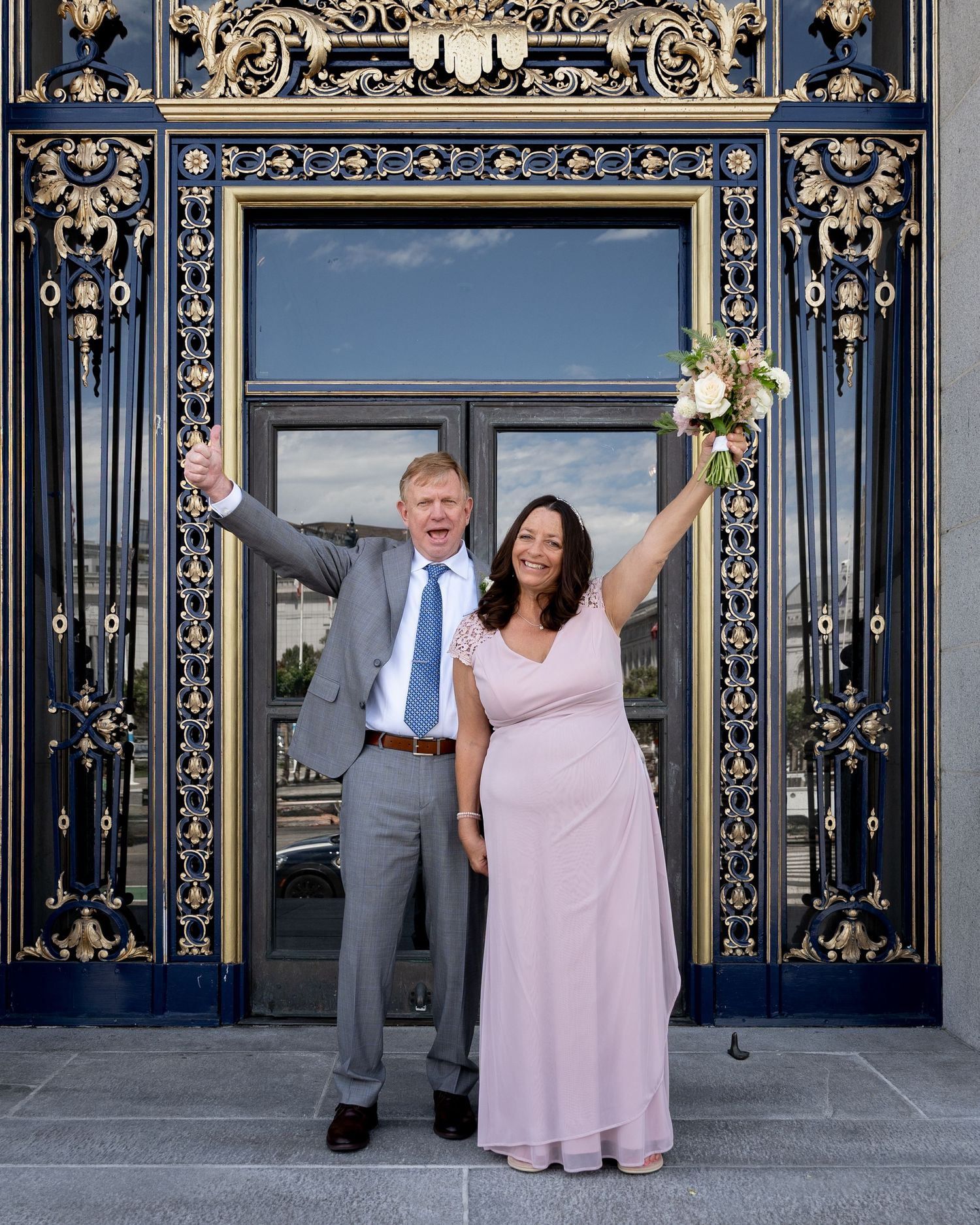 A couple poses together in front of ornate gold and navy gates at San Francisco City Hall.
