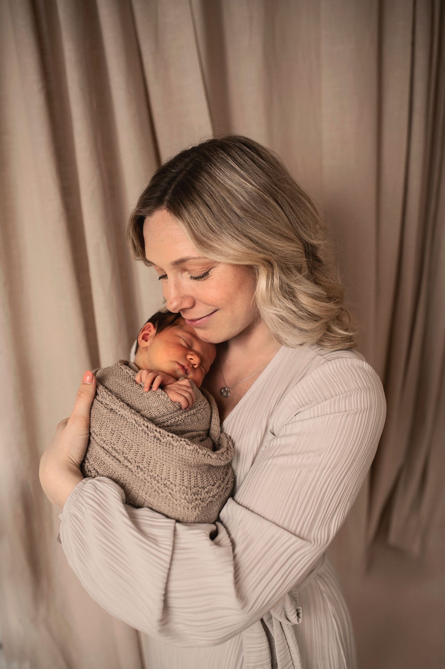 A tender moment captured as a woman in a beige sweater cradles a sleeping newborn wrapped in knit blanket.