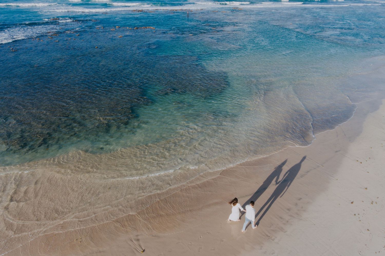 Drone shot of a beach shoreline with clear blue waters and two tiny figures walking.