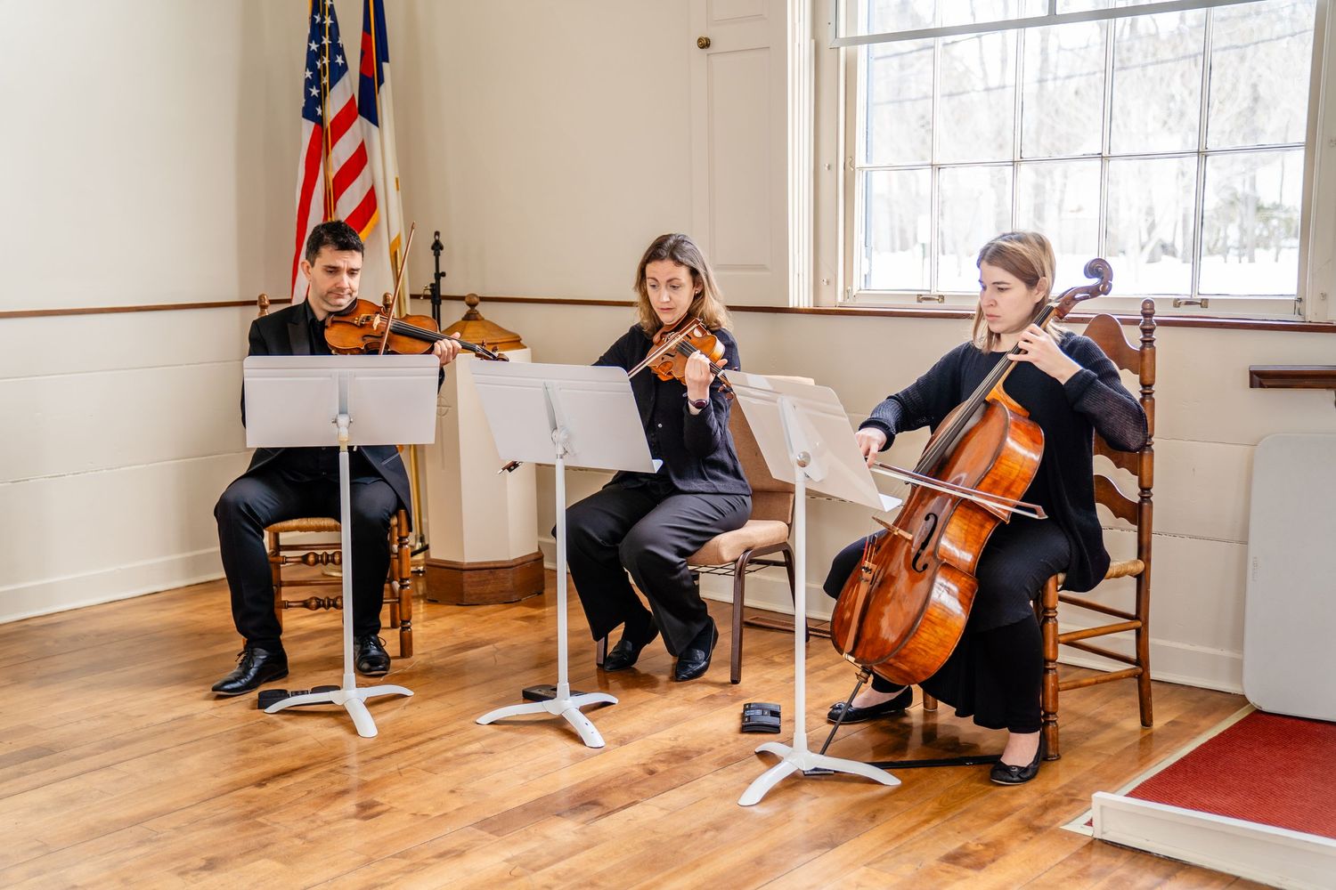 String trio performs on violin, cello, and piano in a bright room with an American flag and hardwood floors.
