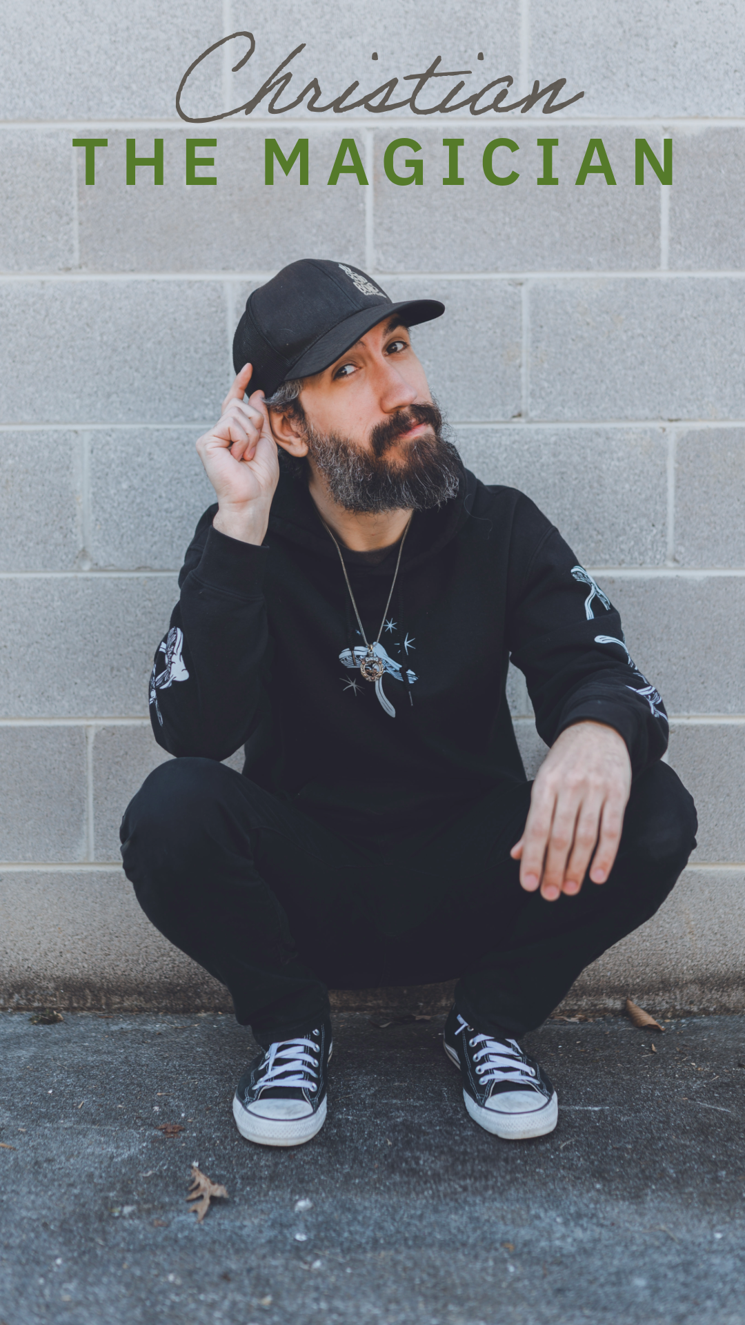 A magician in black attire squats against a concrete wall wearing a baseball cap, sneakers and has decorative sleeve designs.