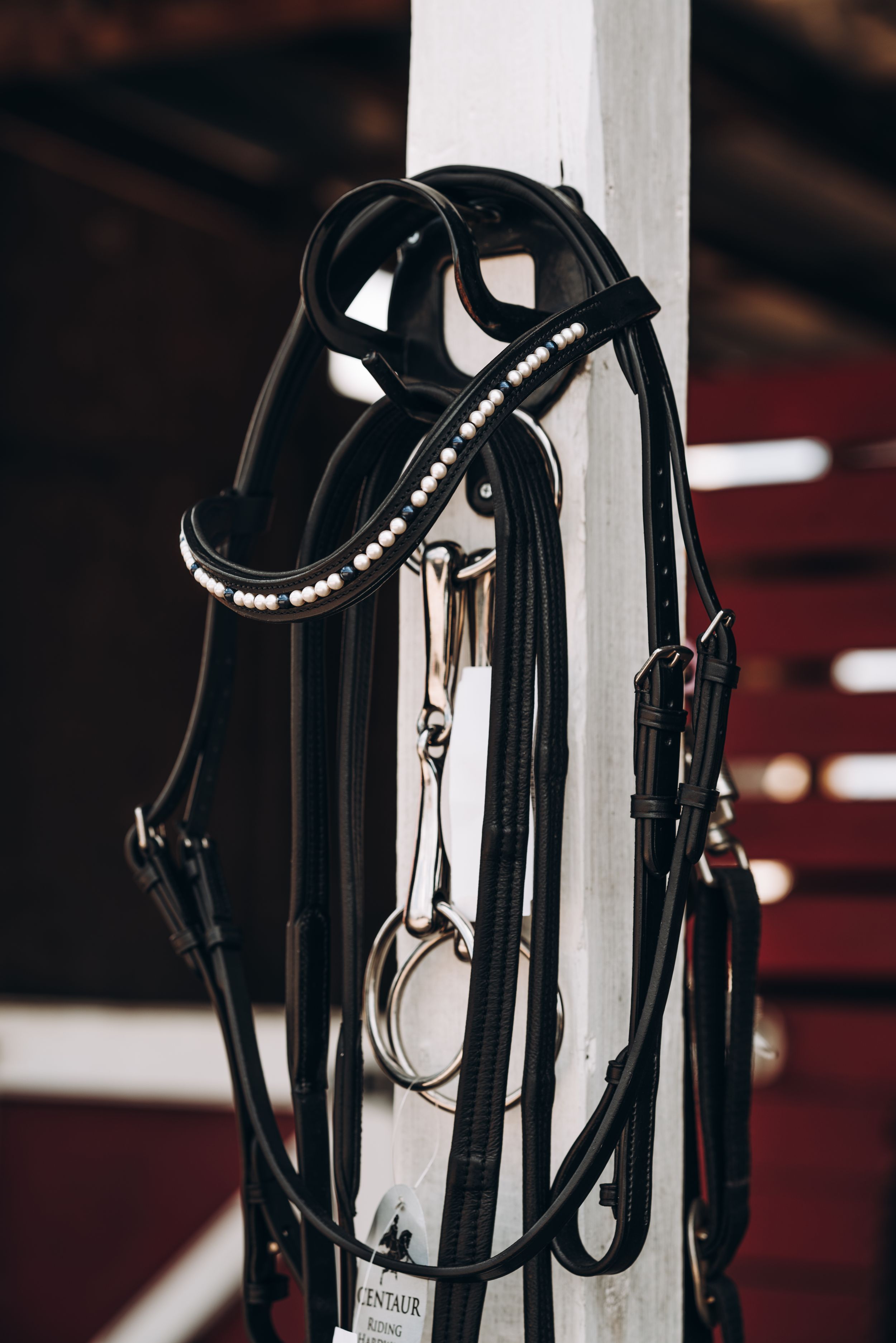 Black leather horse bridle with decorative crystals hanging on white post in stable.
