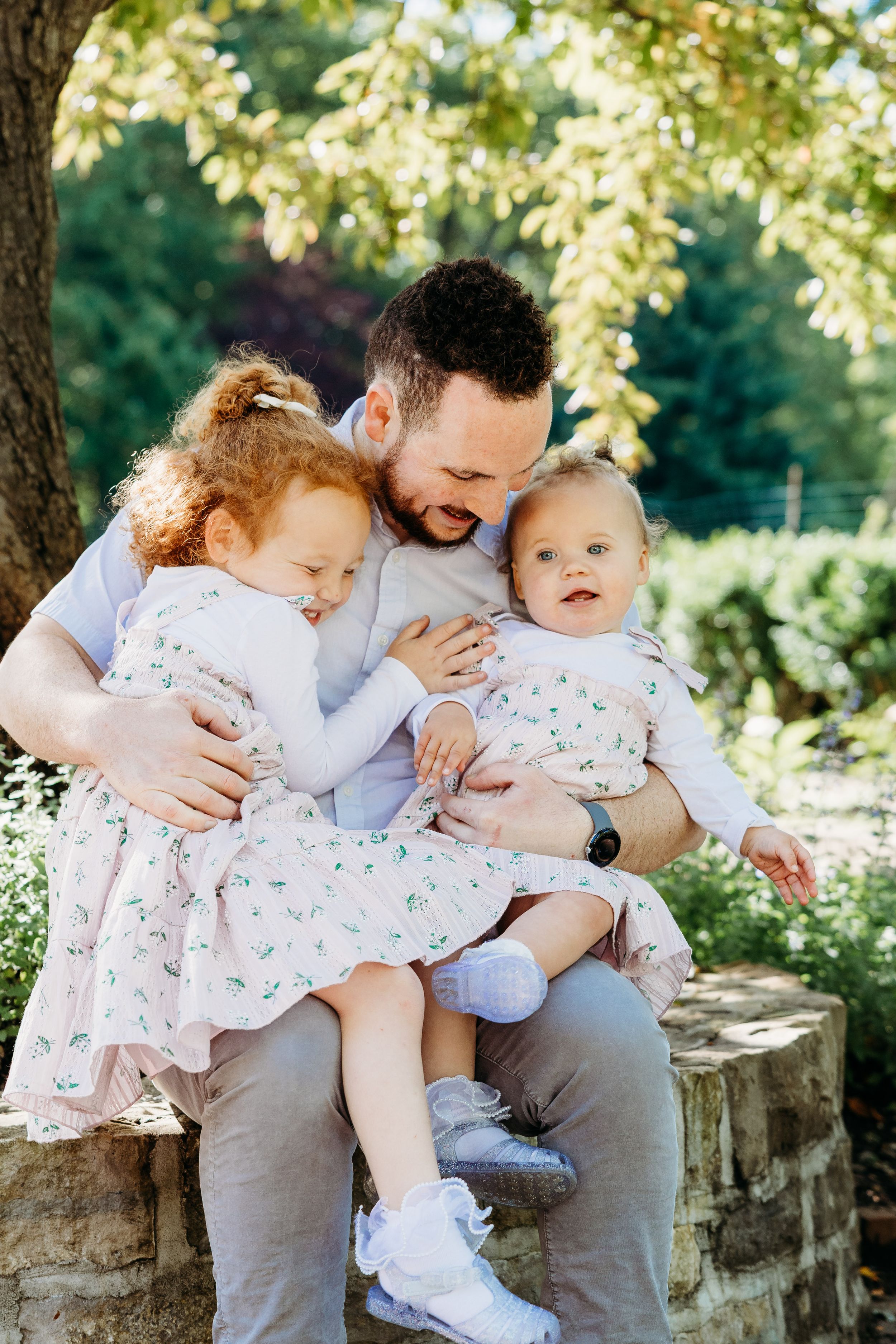 A sweet family moment captured outdoors in dappled sunlight as they share laughs while sitting on a stone wall.