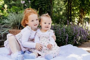 Two young children sit together on a blanket in a garden with blooming purple flowers and greenery.
