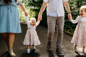 A family walks on a path outdoors while holding hands, with toddler children wearing floral dresses.