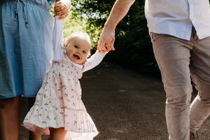 A happy baby in a white patterned dress holds hands with adults while walking outdoors on a sunny day.
