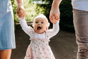 A joyful baby in a patterned dress takes supported steps outdoors while holding hands with adults.