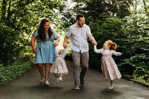 Family of four walking hand in hand along a tree-lined pathway in a lush green park setting.
