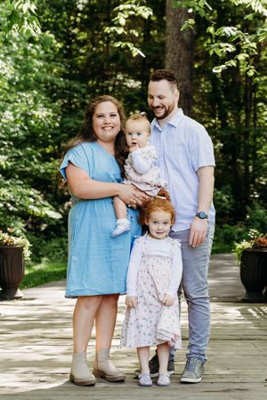 A happy family of four poses together on a wooden deck surrounded by lush summer greenery.