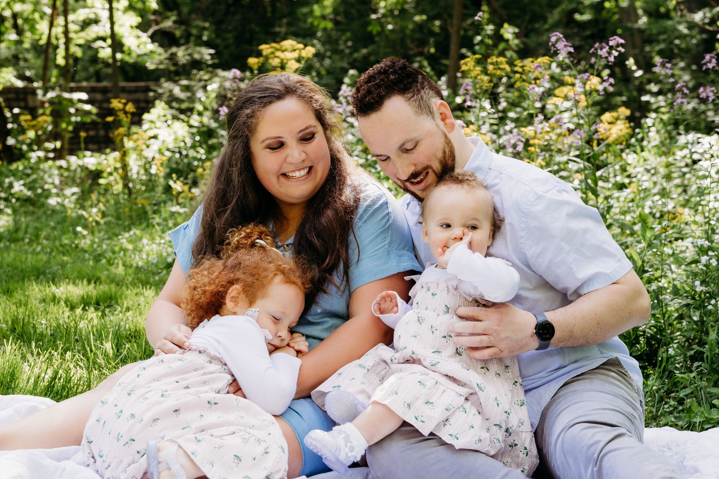 A family sits together on grass surrounded by blooming flowers in a sunny garden setting.