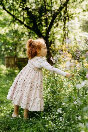 A child in a floral dress reaches out to touch blooming wildflowers in a sunlit garden with trees in the background.