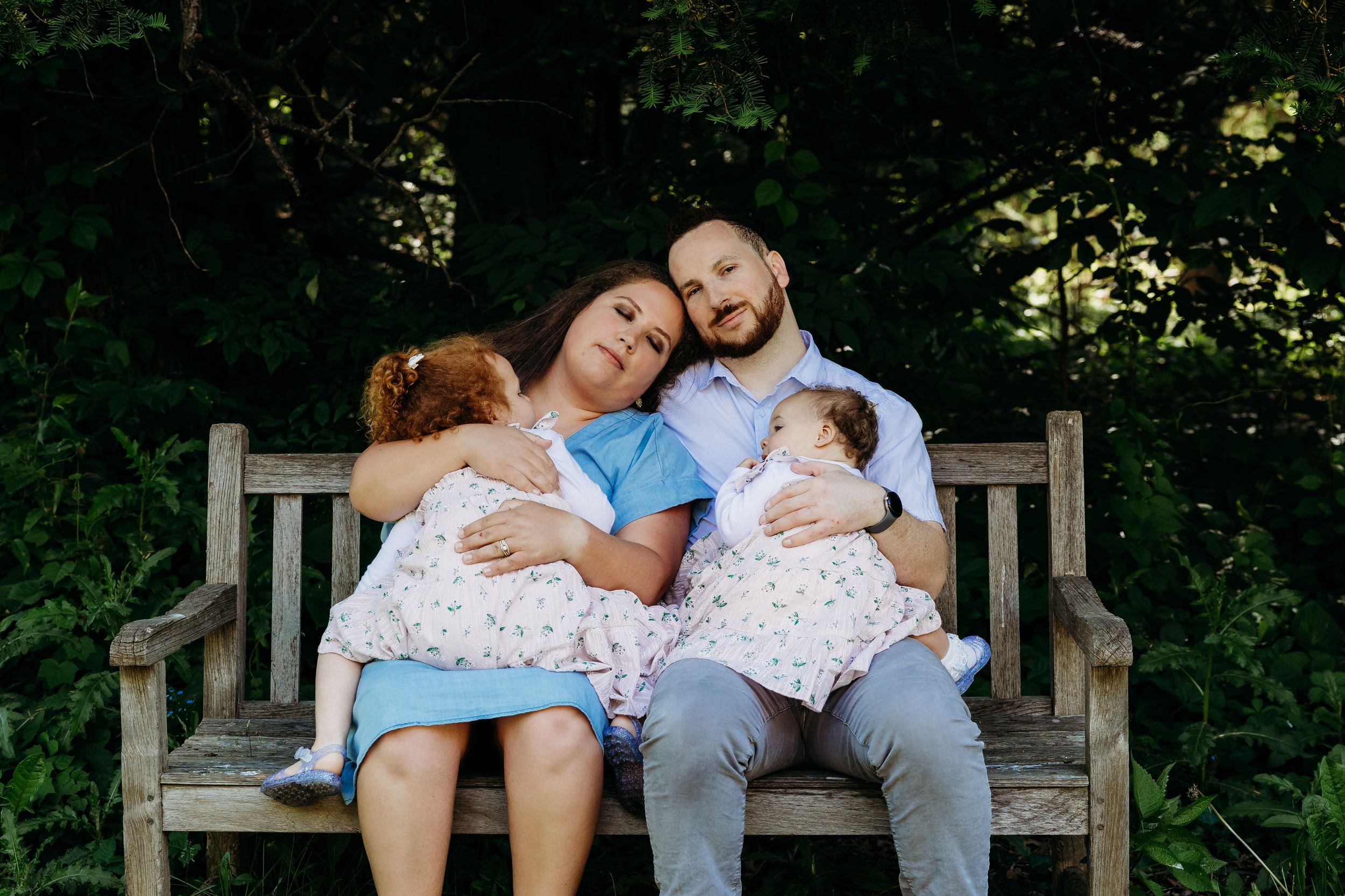 Two adults and two small children resting together on a wooden garden bench in a peaceful outdoor setting.