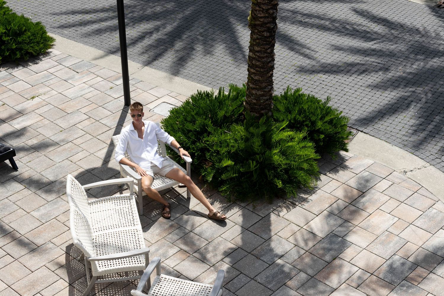 Man in white outfit relaxing on wicker chair near palm tree on sunny outdoor patio with brick pavers.