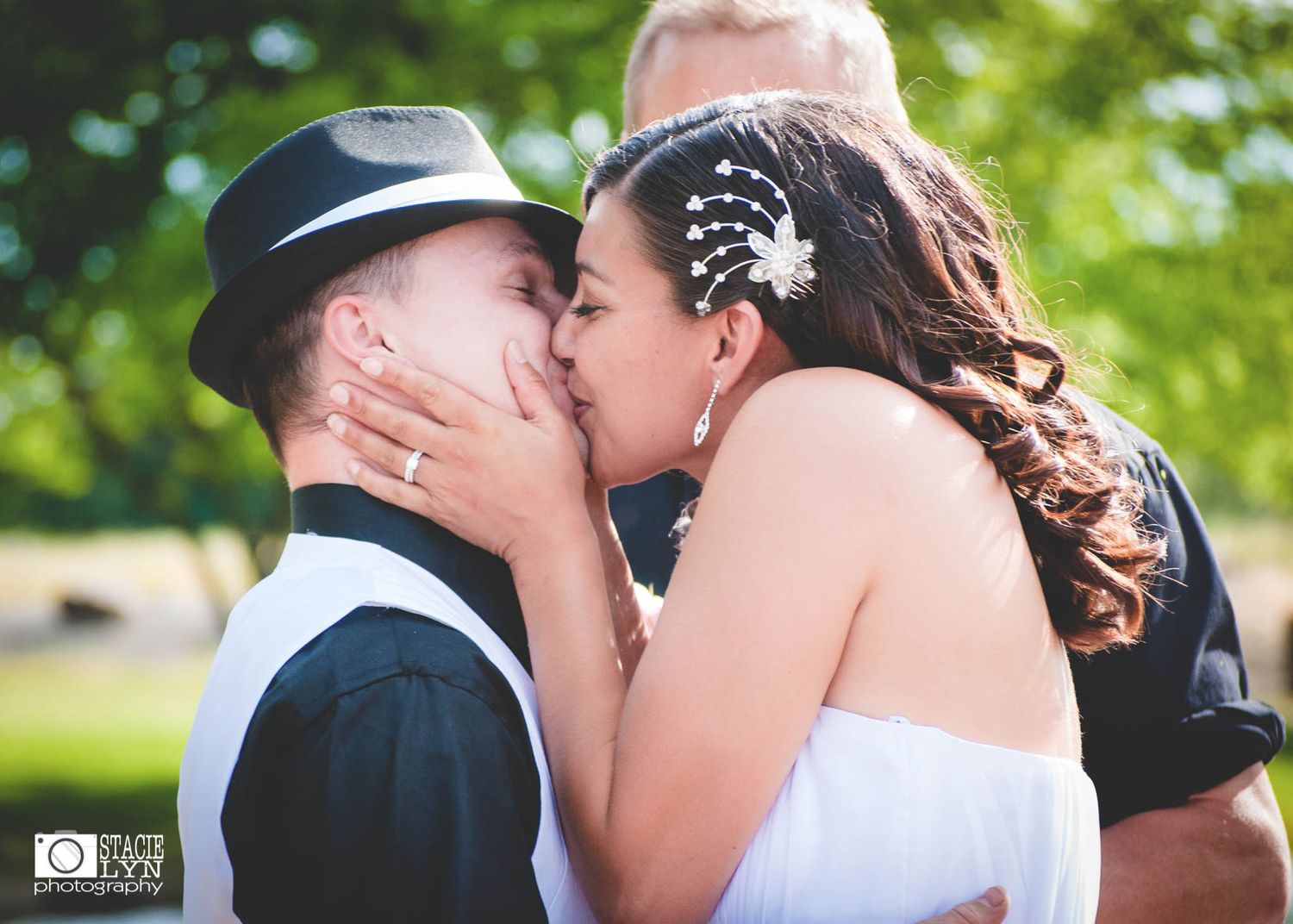 A couple shares a tender kiss outdoors in wedding attire on a sunny day with greenery in the background.