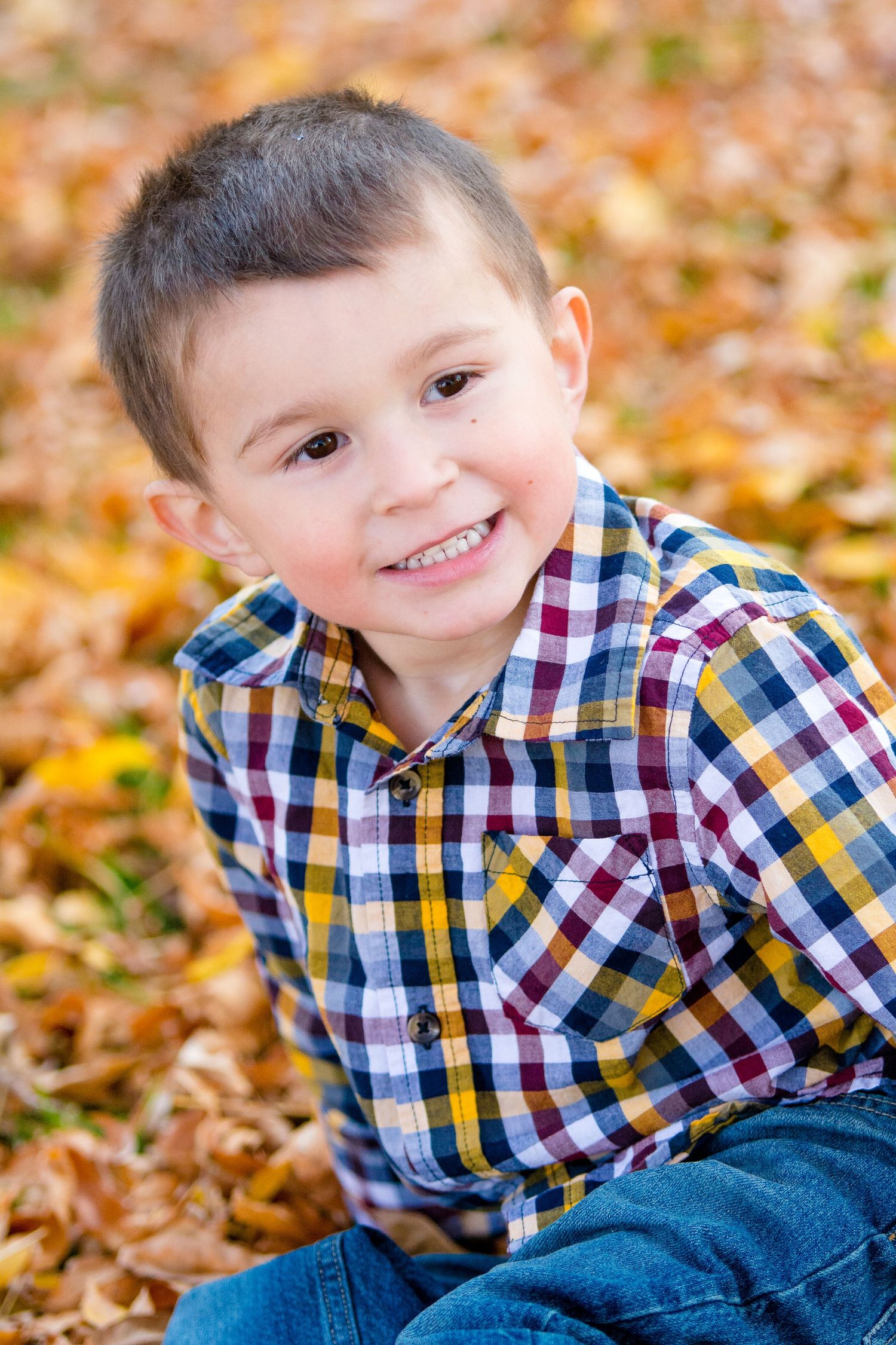 Young child wearing a colorful plaid shirt smiles brightly while sitting among fallen autumn leaves.