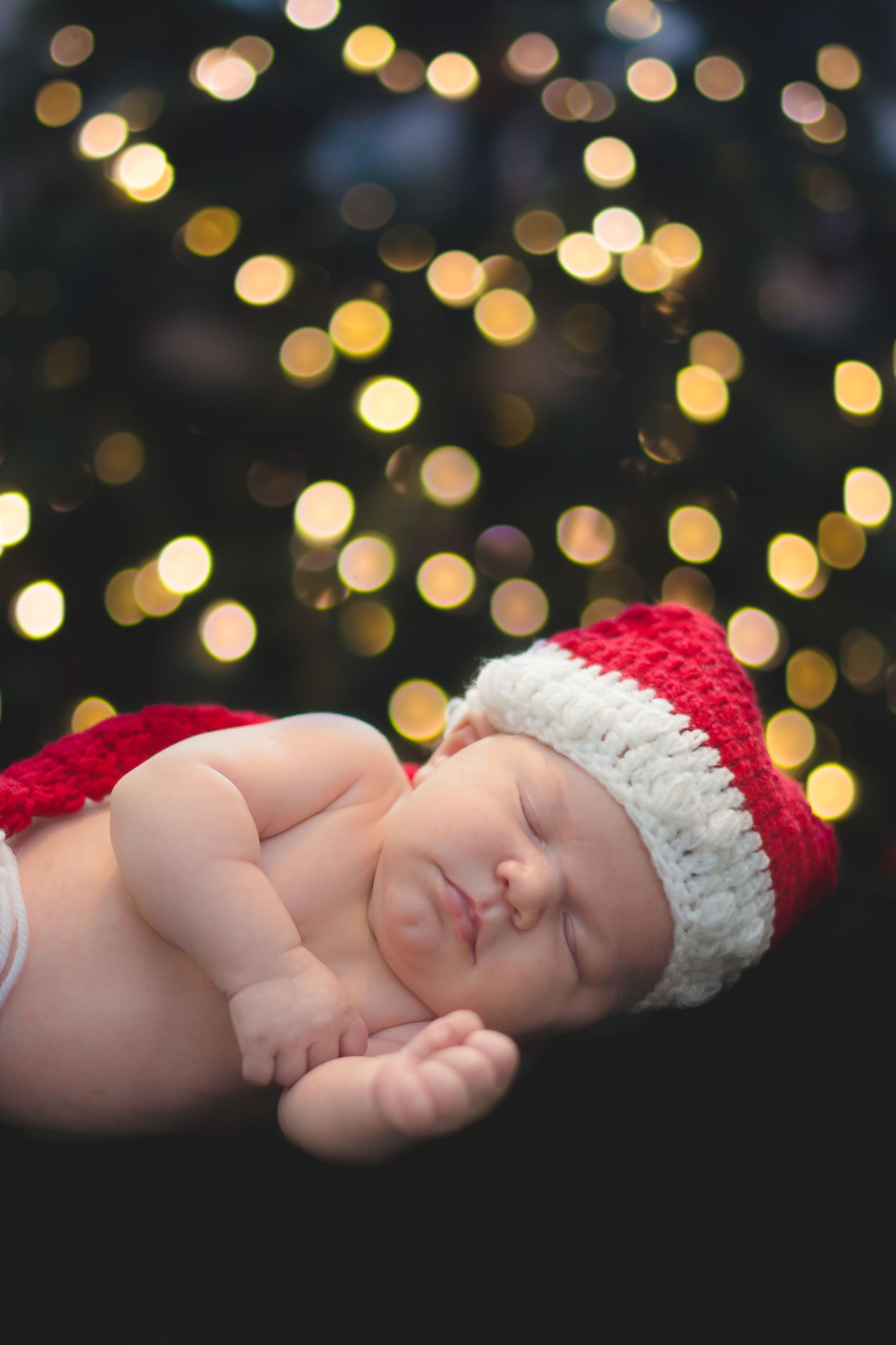 Sleeping newborn wearing red and white knit holiday hat with glowing bokeh lights in dark background.