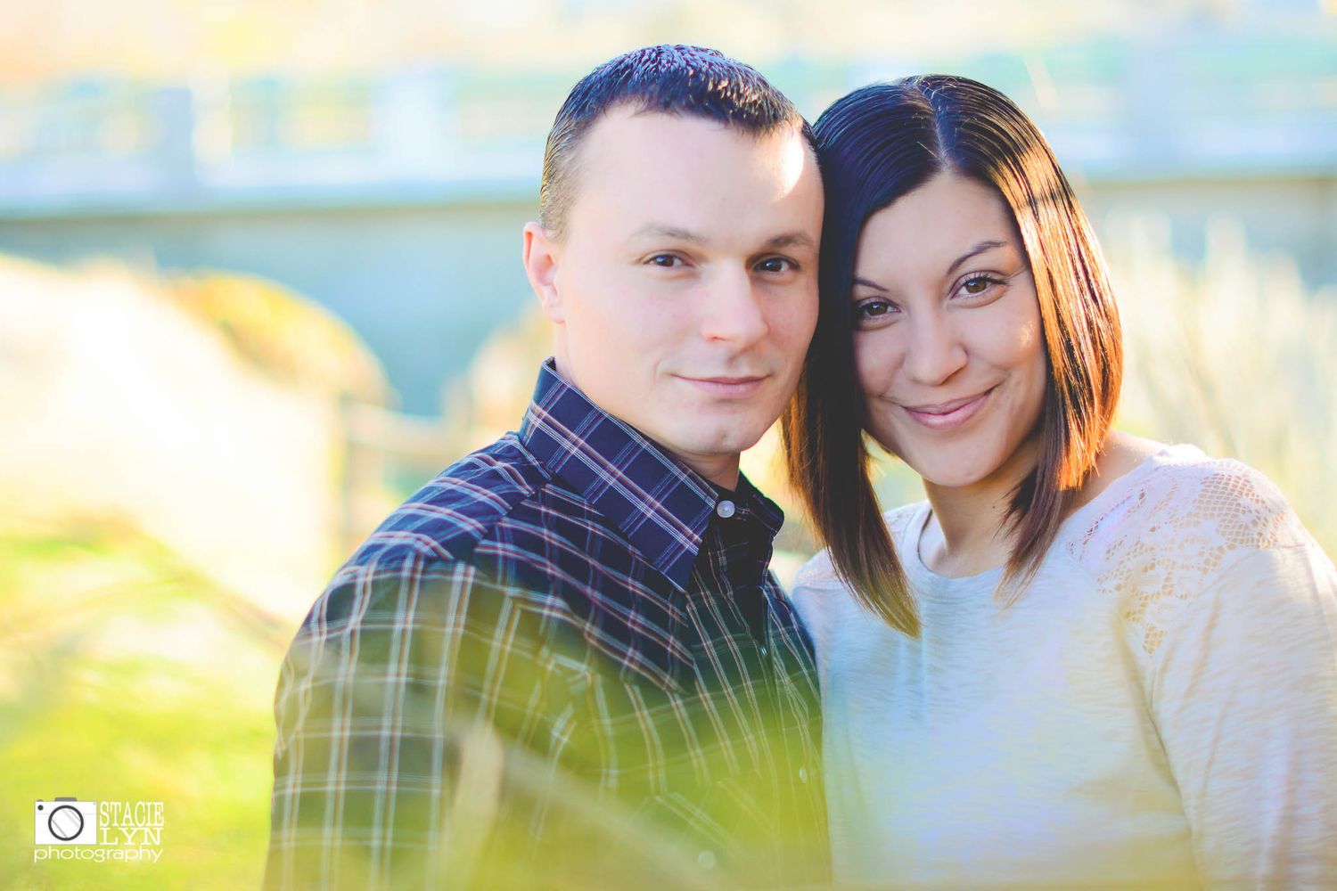 A couple smiles together outdoors in natural lighting with blurred greenery and sunlight in the foreground.