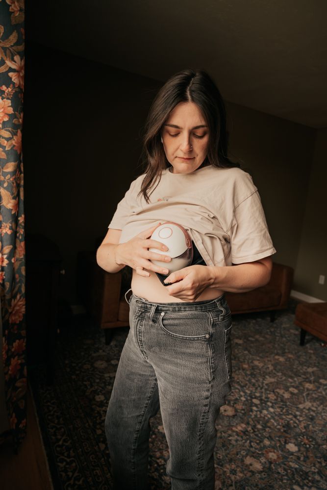 Person in beige t-shirt and gray jeans looking down while standing in a dimly lit room with floral curtains.