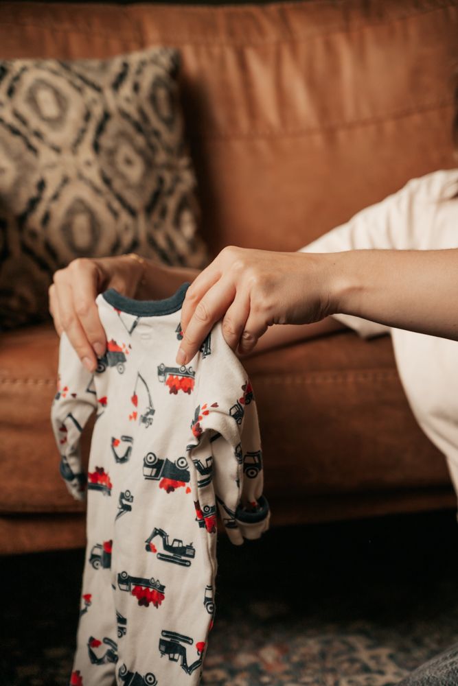 Hands folding and organizing small gray baby onesies with red and blue trucks with hearts  patterns on a brown couch.