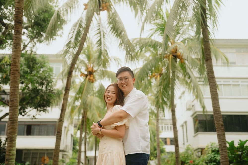 A couple embraces while standing among tall palm trees outside a white building on a tropical day.