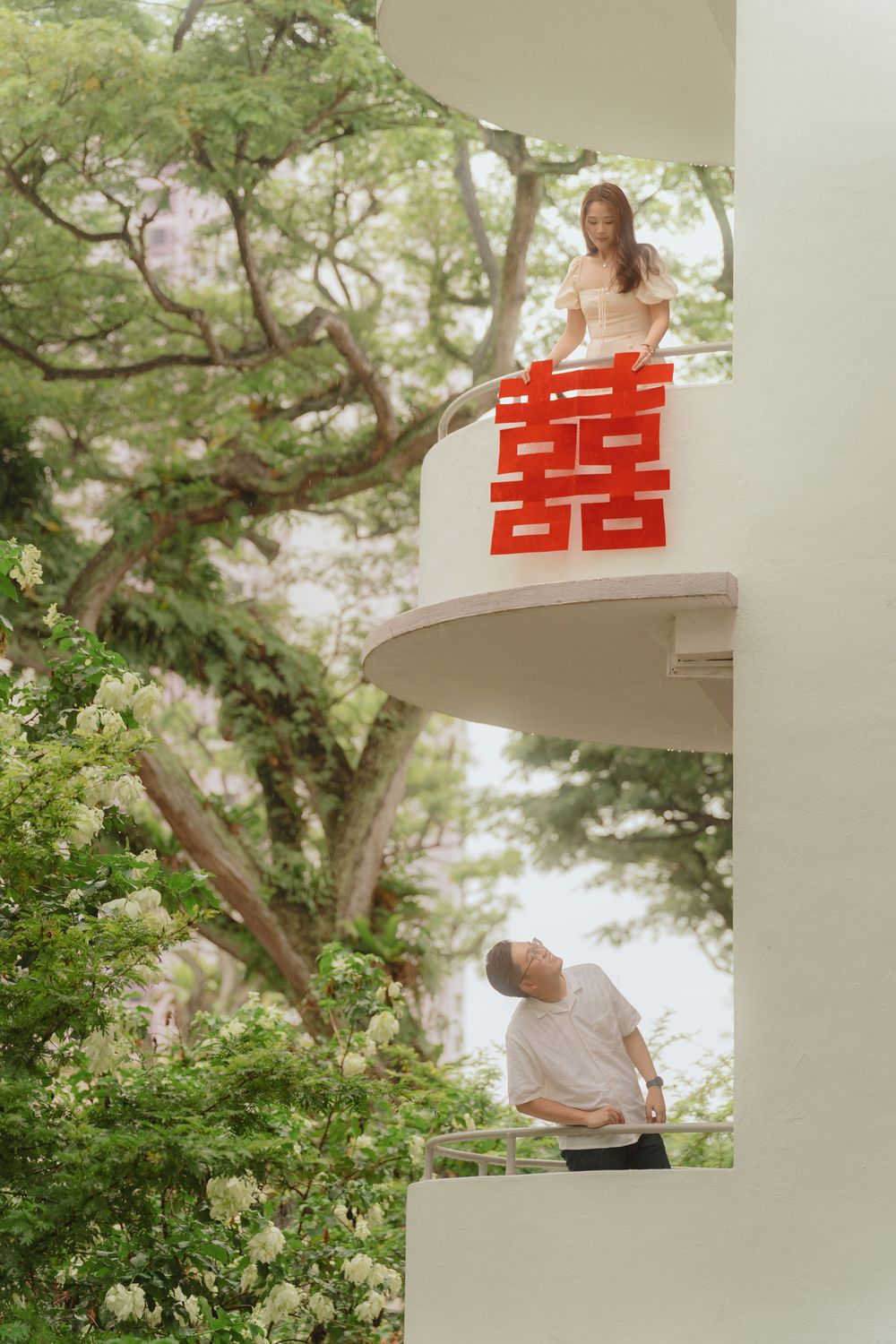 Two people in traditional wedding attire pose between levels of a white building with red double happiness symbol.
