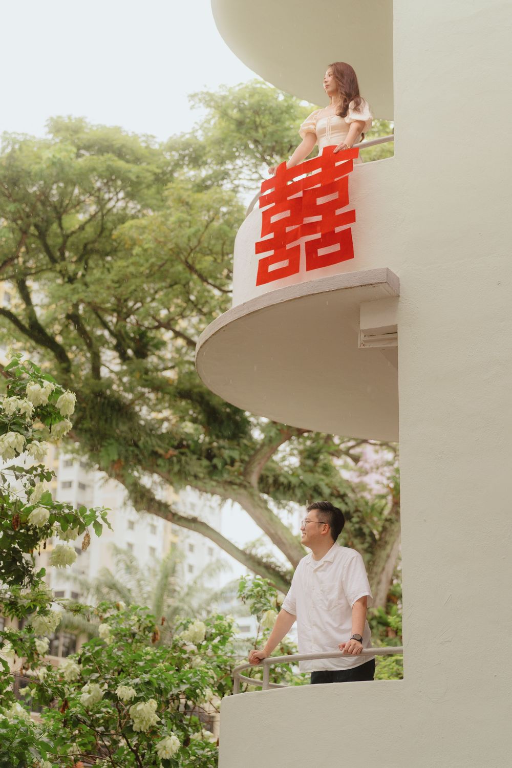 Two people stand on different levels of a white balcony with a red double happiness symbol and lush greenery in background.