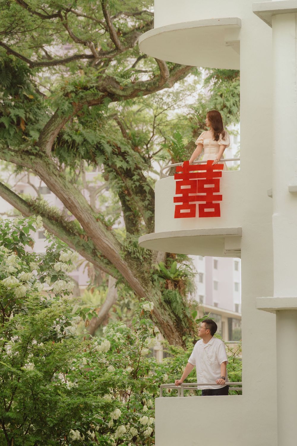 Couple on different levels of white balcony with red Chinese double happiness symbol and lush greenery in background.