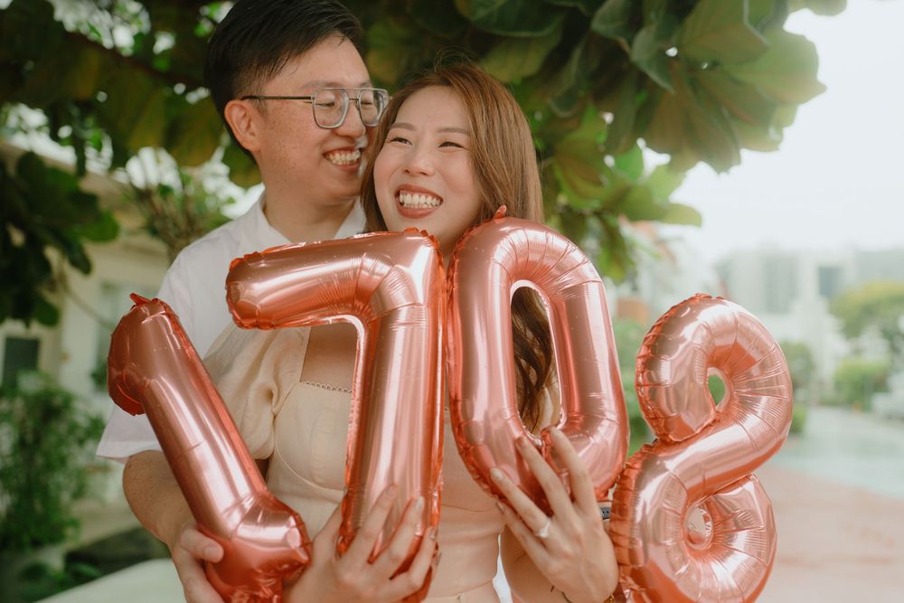 Couple stands together outdoors holding rose gold number balloons spelling 708 against green foliage background.