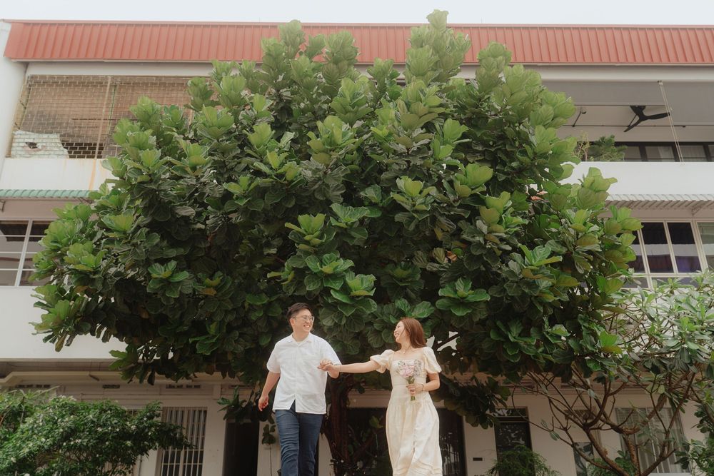 A couple stands beside a large lush green tree in front of a building with a terracotta roof.