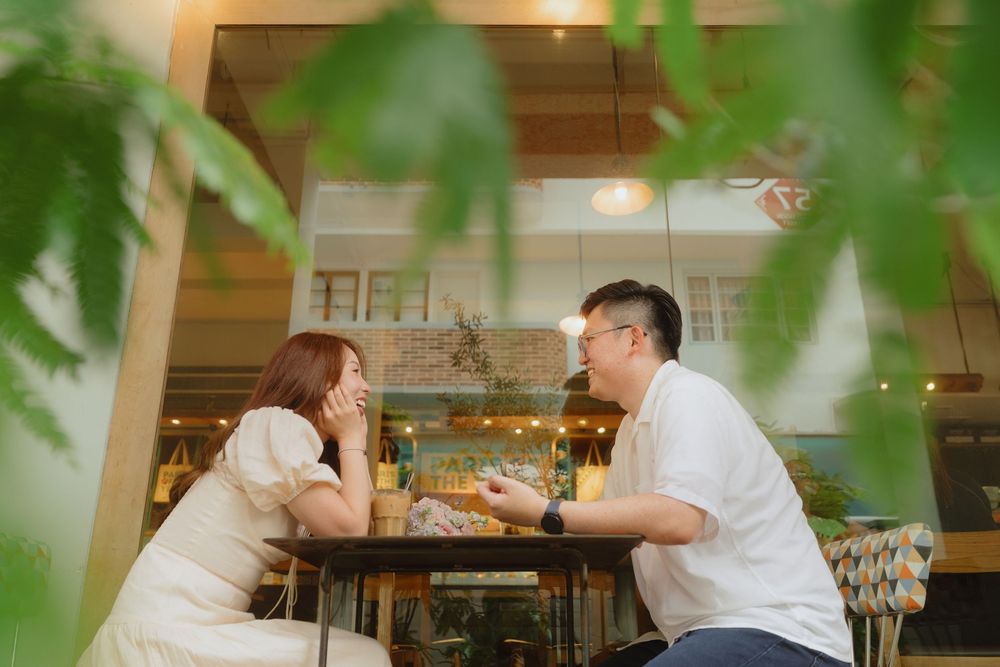 Two people share an intimate conversation at a restaurant table framed by green leaves in the foreground.