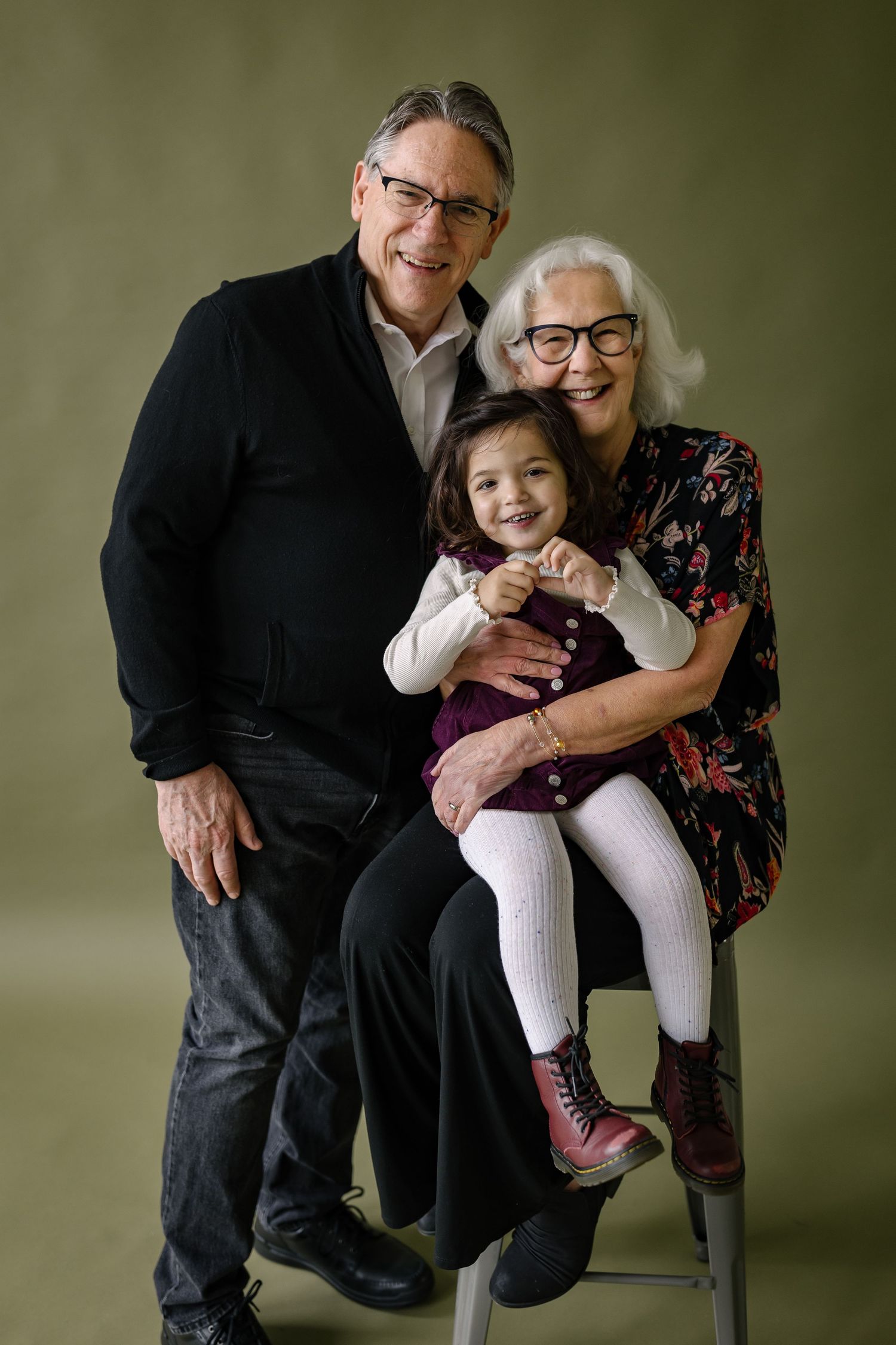 A young child wearing pink boots sits between two adults posing together against a green studio backdrop.