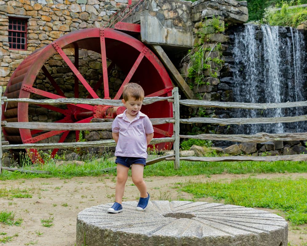 A toddler boy plays candidly at Wayside Inn Grist Mill during a family photo session in Sudbury with Lisa West Photography outside Boston.