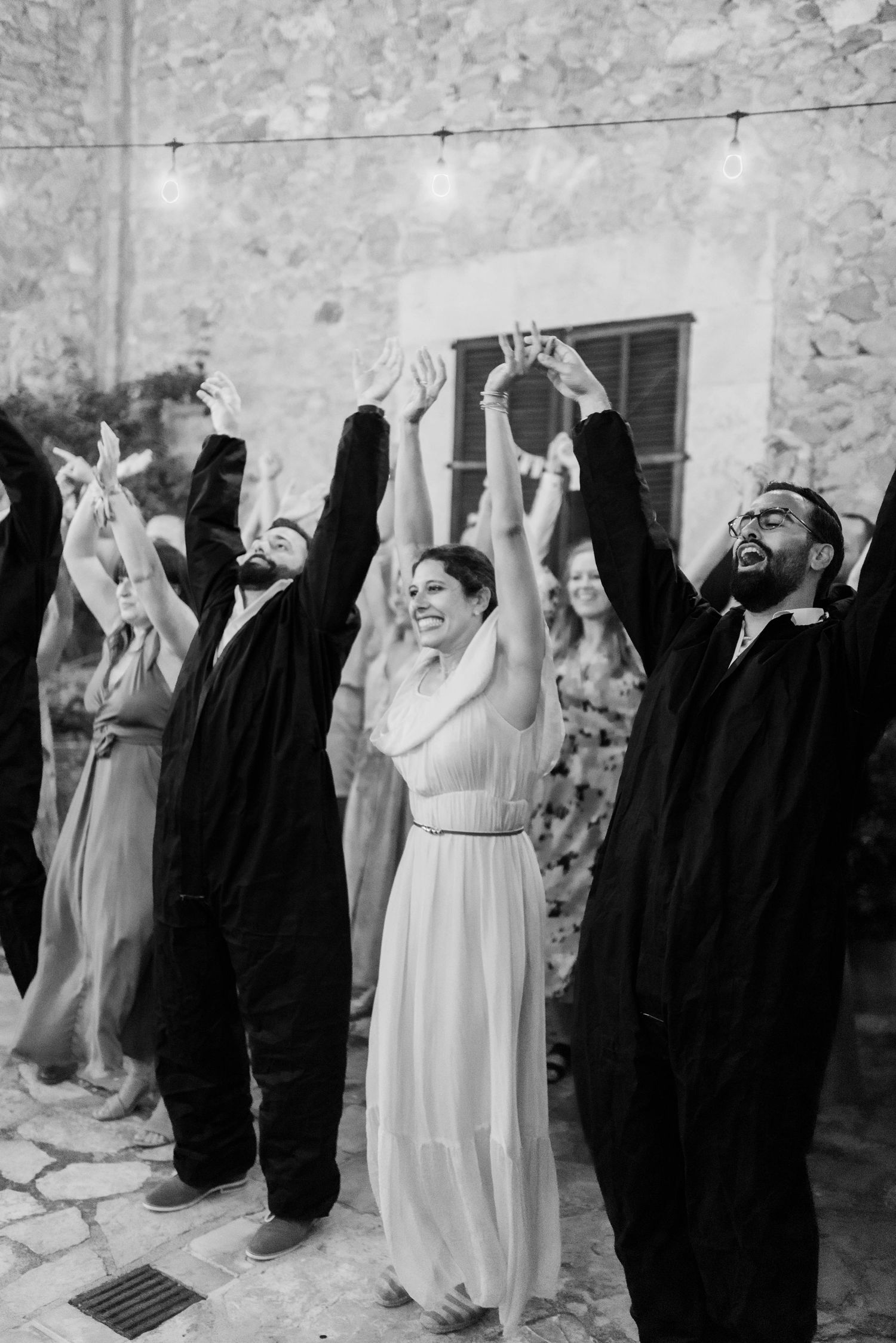 Eine Gruppe von Feiernden in schwarz-weißer Kleidung hebt während eines festlichen Moments bei einer Feier im Freien freudig die Arme.