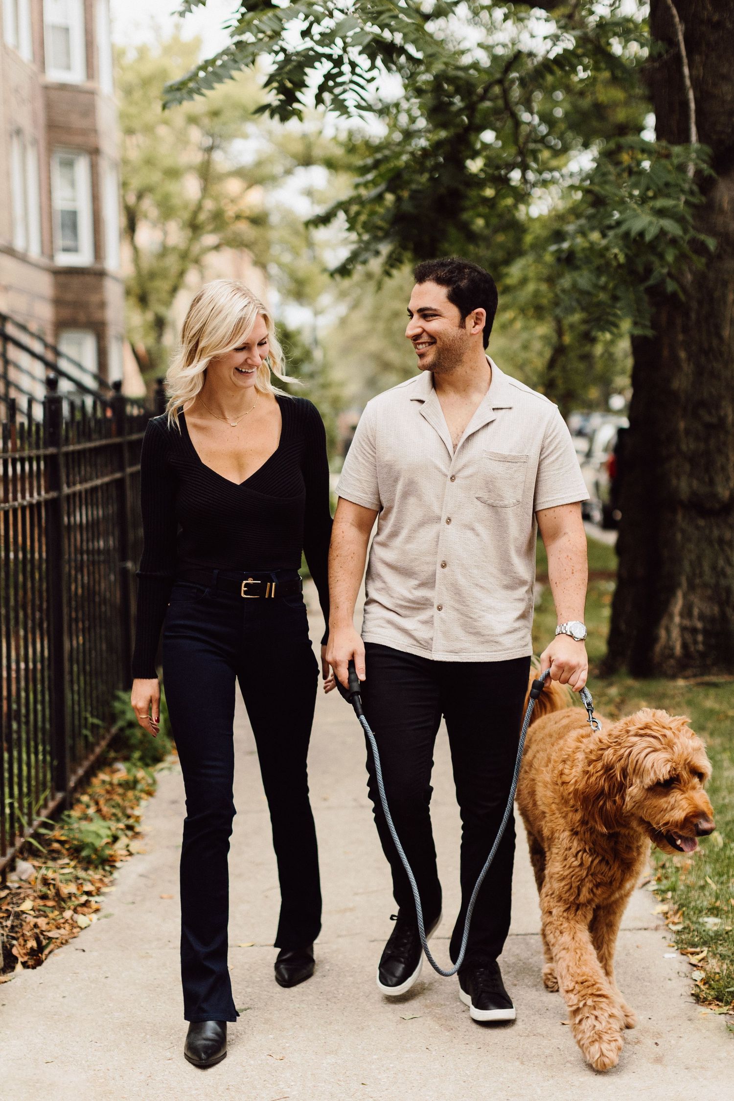 A couple walking their goldendoodle dog on a tree-lined city street.