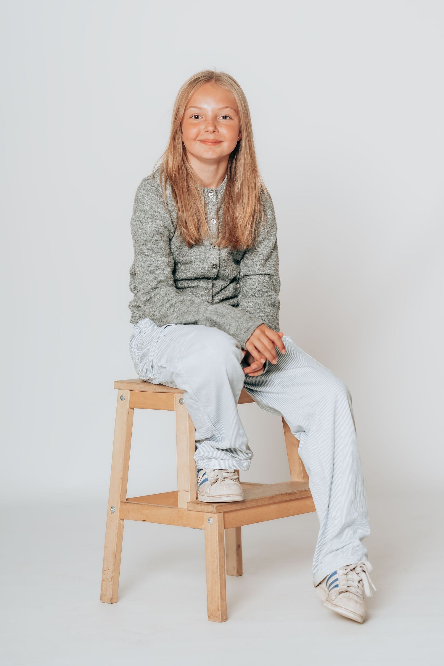 Studieportræt af et smilende barn siddende på en træskammel i et lyst fotostudie.