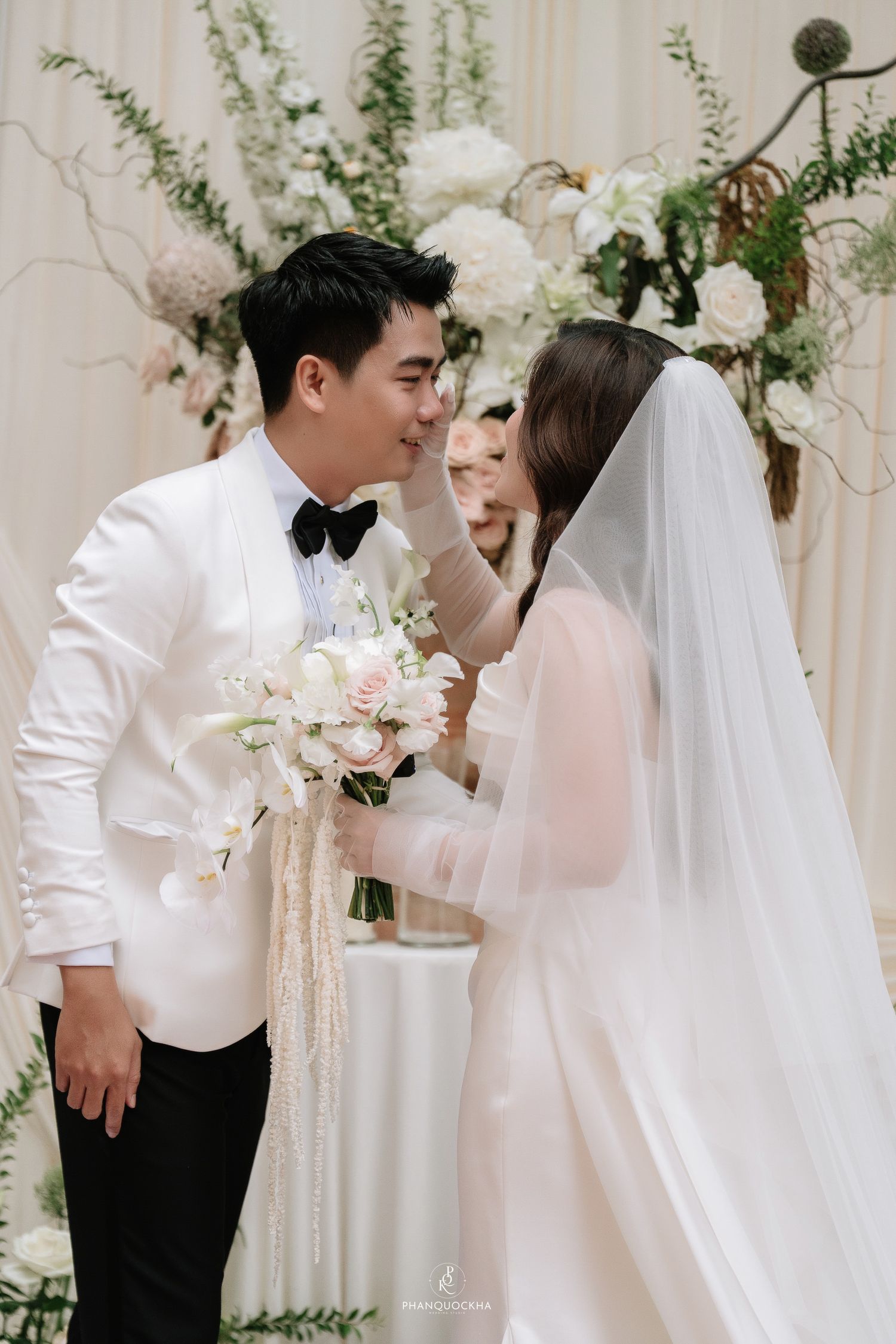 Groom in white tuxedo leans in to kiss bride holding white bouquet at elegant floral wedding altar.